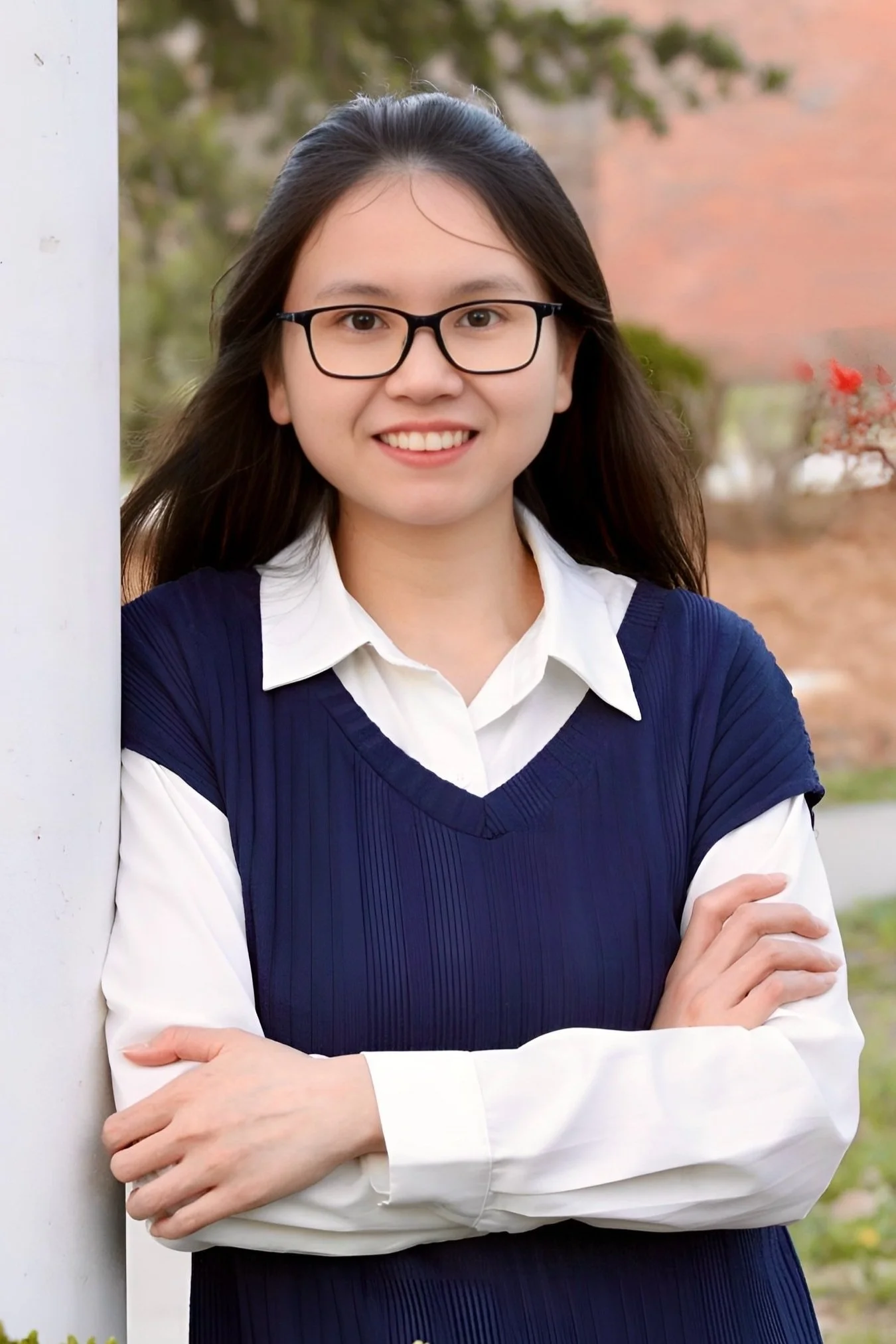 A person with long hair, wearing glasses, a navy dress, and a white shirt stands outdoors, smiling and leaning against a white pillar.
