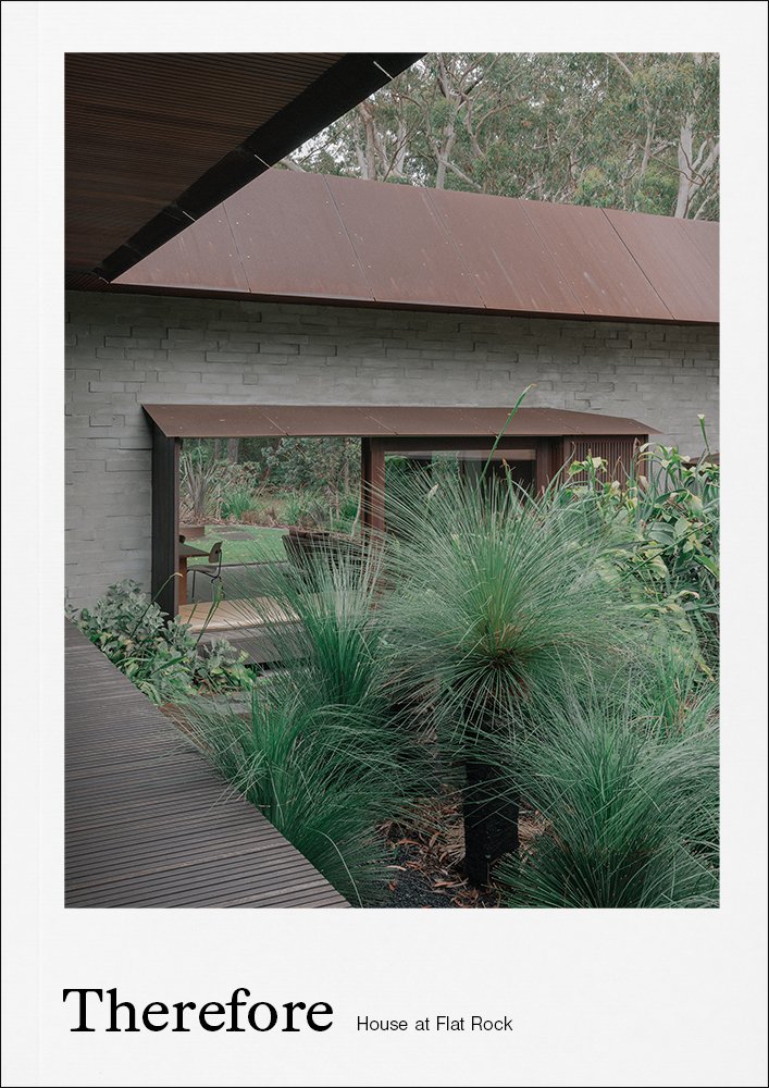 A modern house with gray brick walls, a rust-colored metal roof, and a large window with wooden framing surrounded by lush green plants and trees.