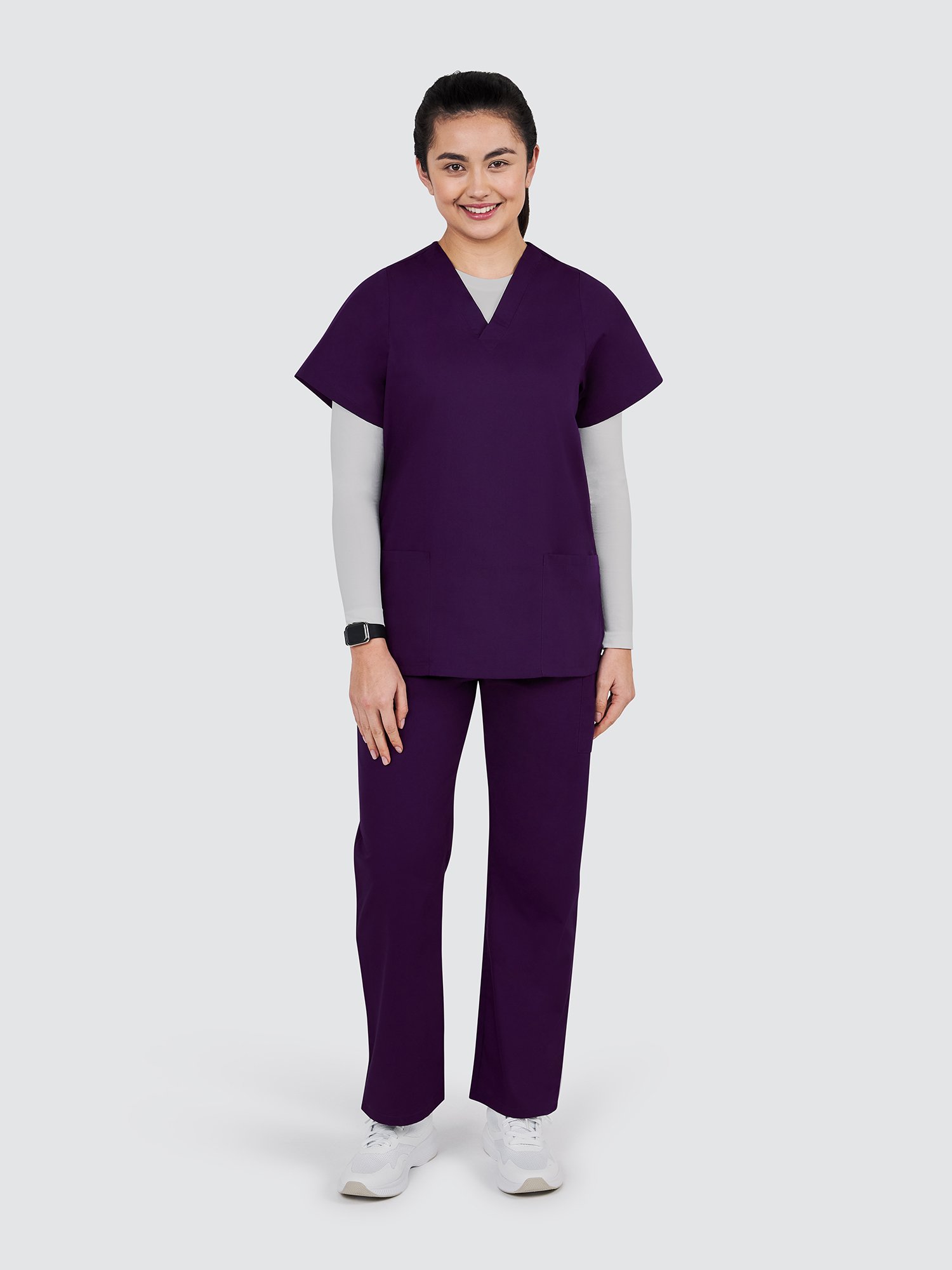 A person stands smiling, wearing purple scrubs and white sneakers against a plain background. Their hair is neatly tied back, and they wear a light gray long-sleeve shirt under the scrubs.