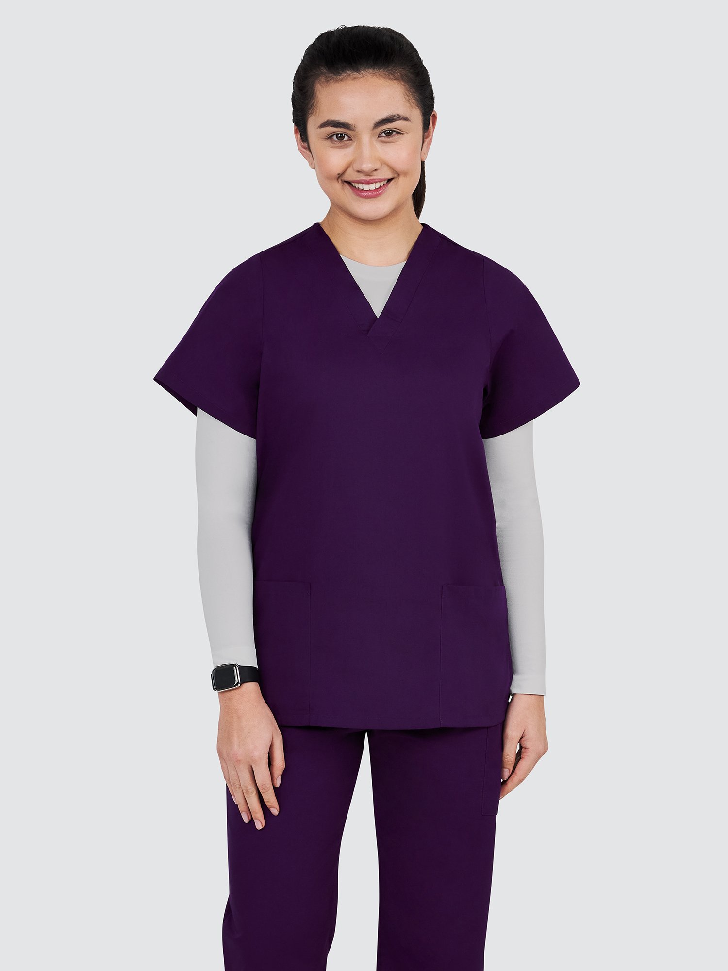 A person stands smiling, wearing purple scrubs and white sneakers against a plain background. Their hair is neatly tied back, and they wear a light gray long-sleeve shirt under the scrubs.