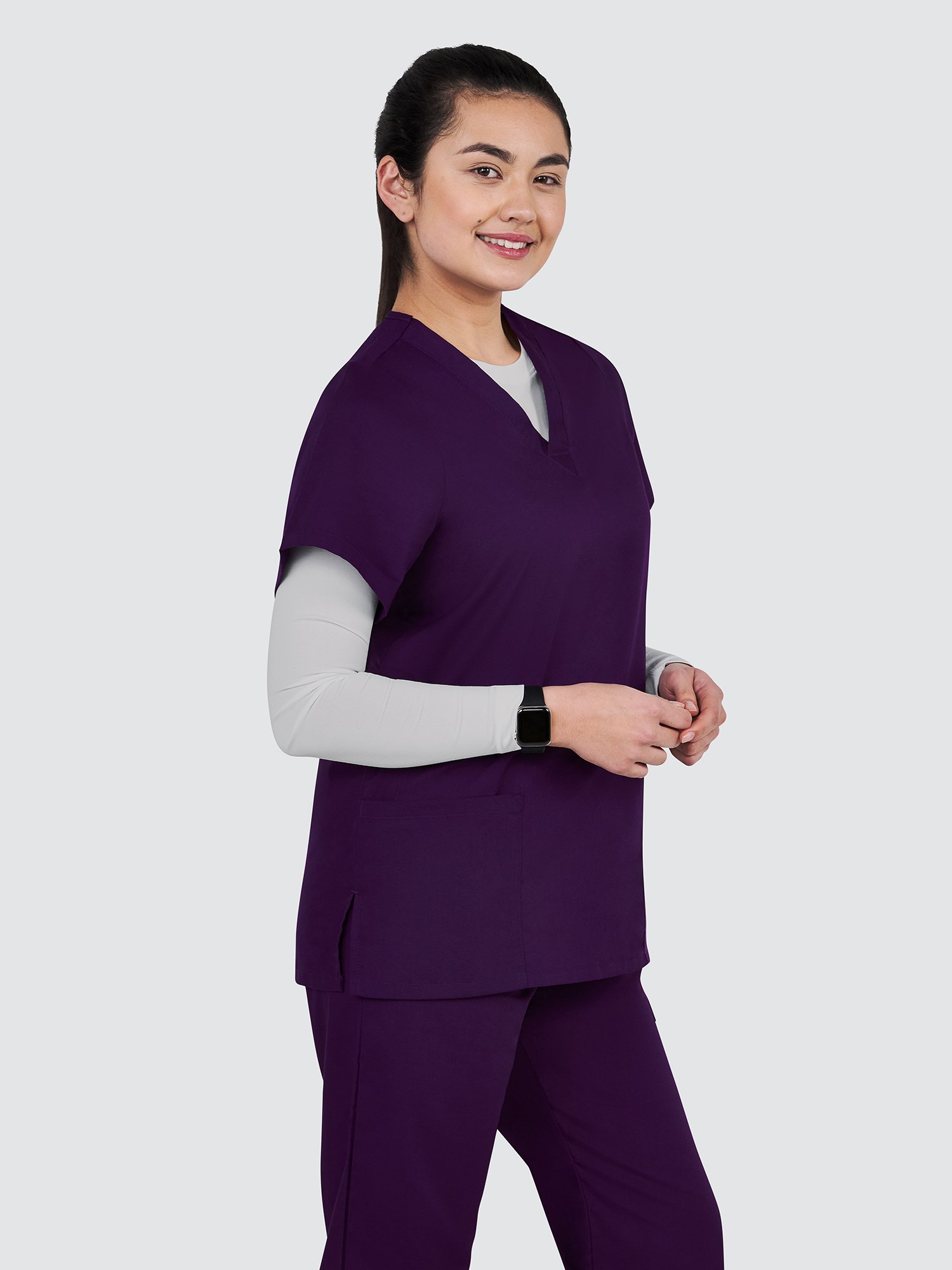 A person wearing a purple scrub top and pants with a light gray undershirt stands smiling against a plain background. They are also wearing a smartwatch on their left wrist.