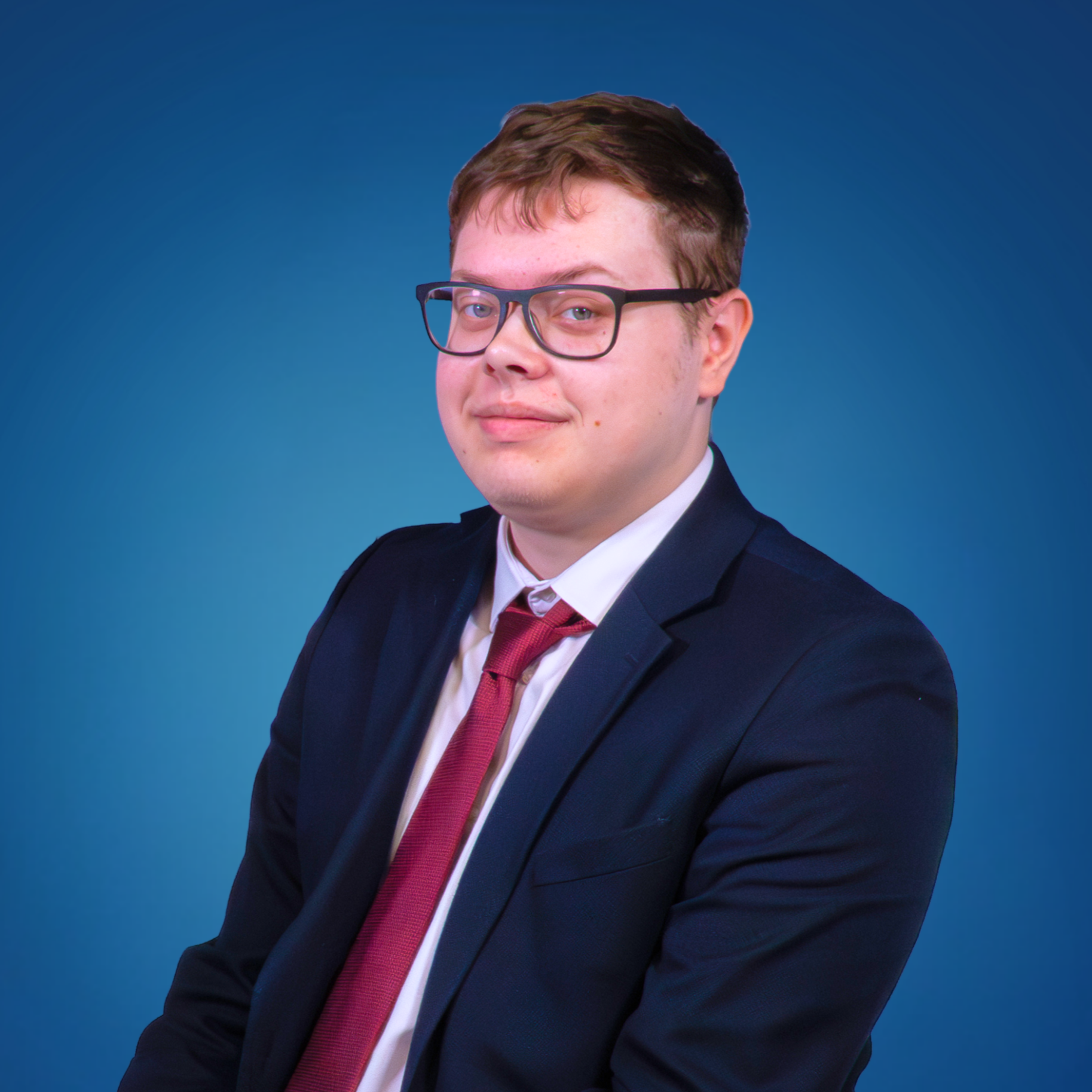 A young man with glasses wearing a navy suit, white shirt, and maroon tie, posing against a blue background.