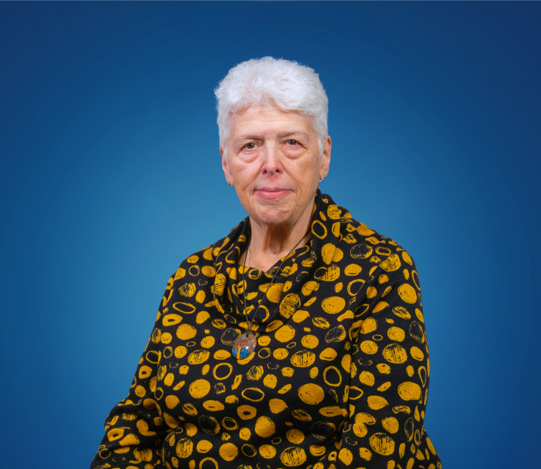 An elderly woman with short white hair, wearing a black and yellow patterned top, with a necklace featuring a colorful circular pendant, poses against a blue background.