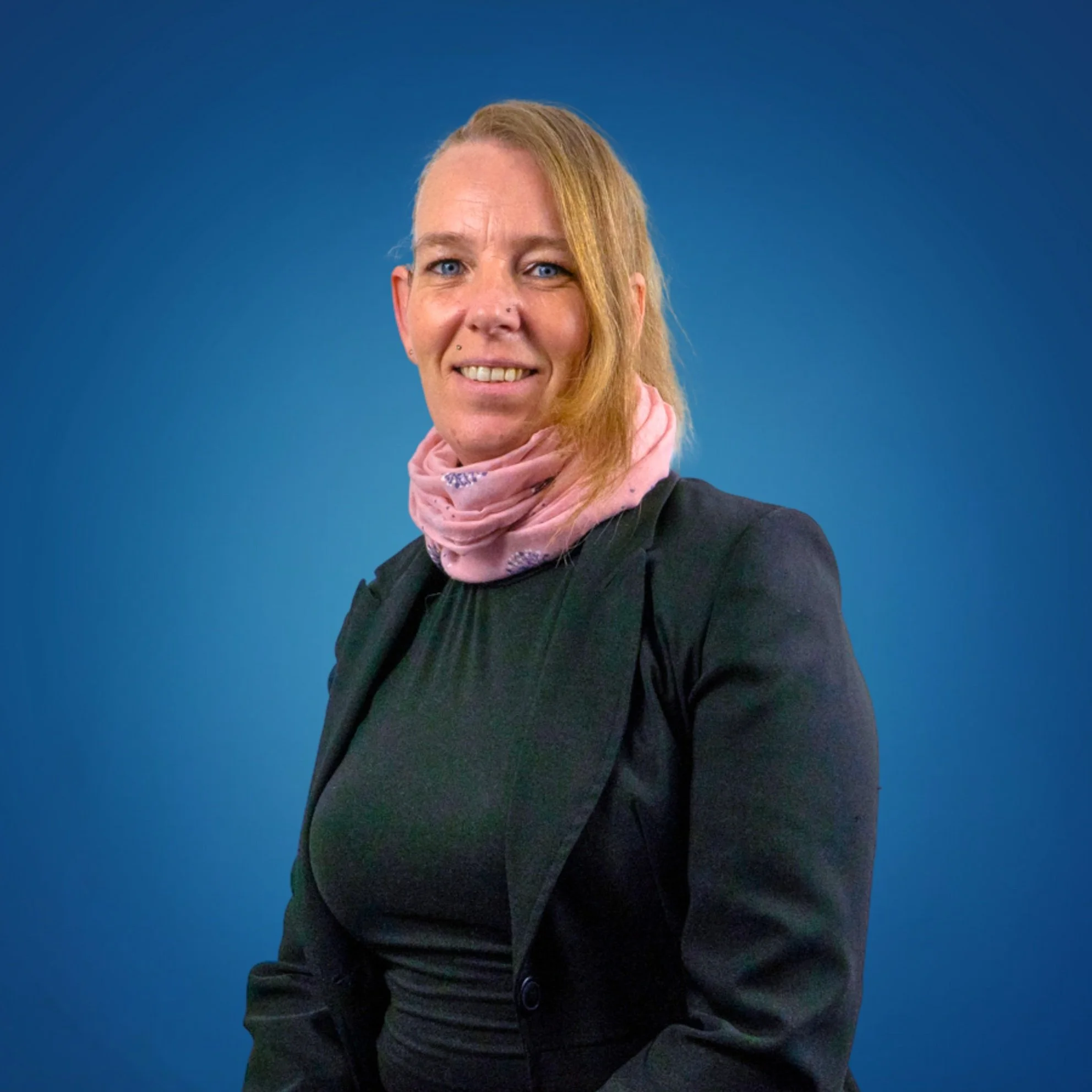 Portrait of a woman with blonde hair, wearing a black blazer and pink scarf, smiling against a blue background.