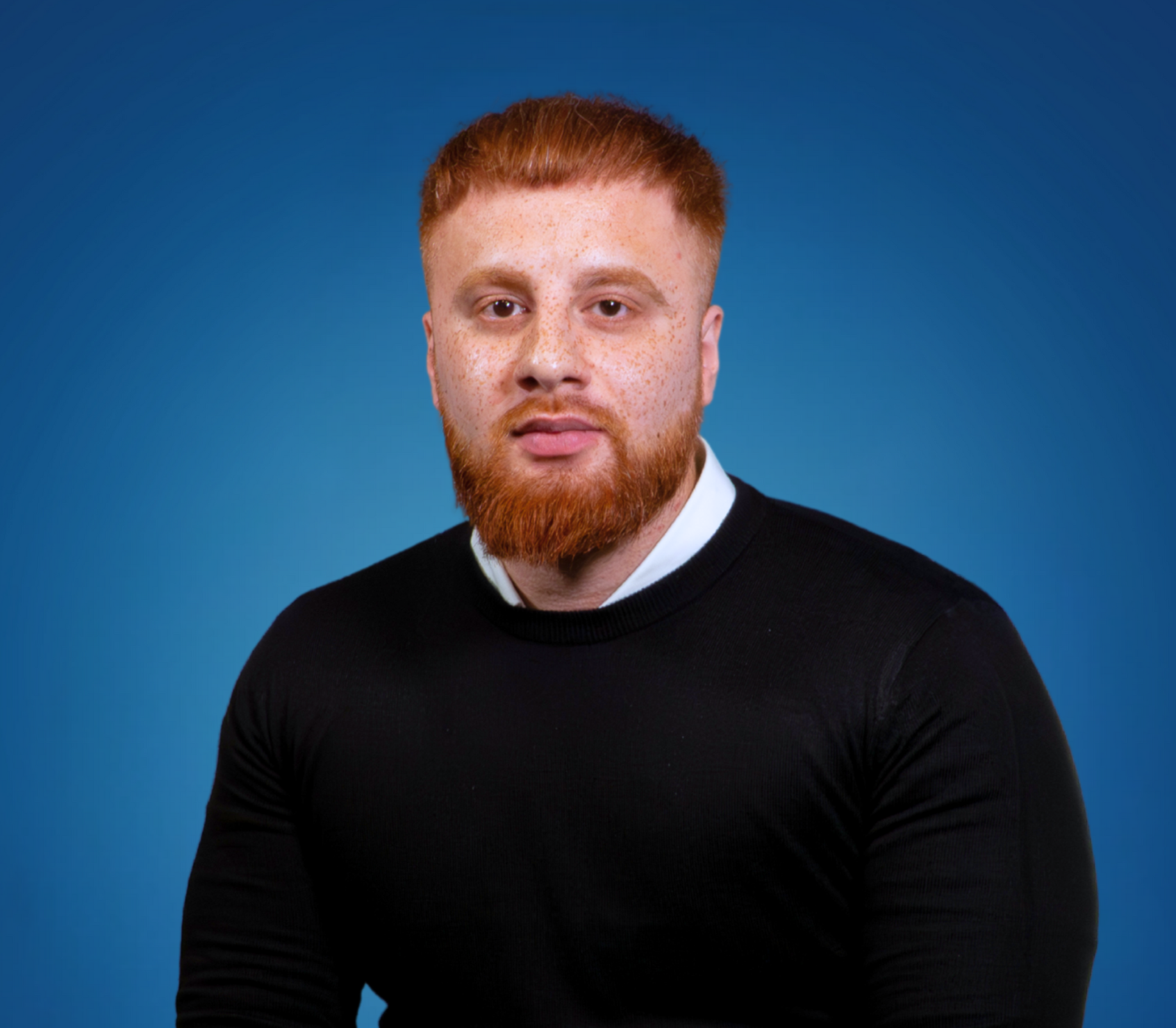 A young man with red hair and beard wearing a black sweater over a white collared shirt against a blue background.