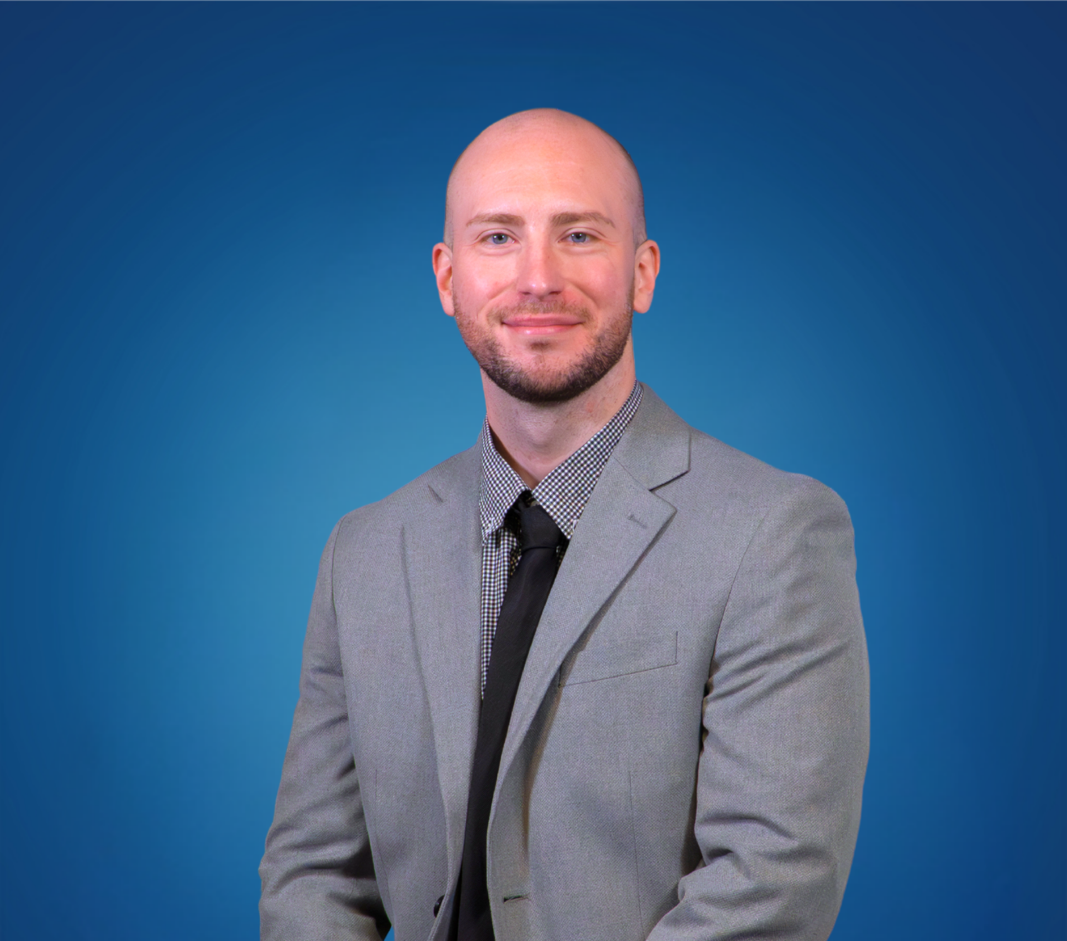 A man with a bald head, beard, and light skin wearing a gray suit, patterned shirt, and black tie, smiling against a blue background.