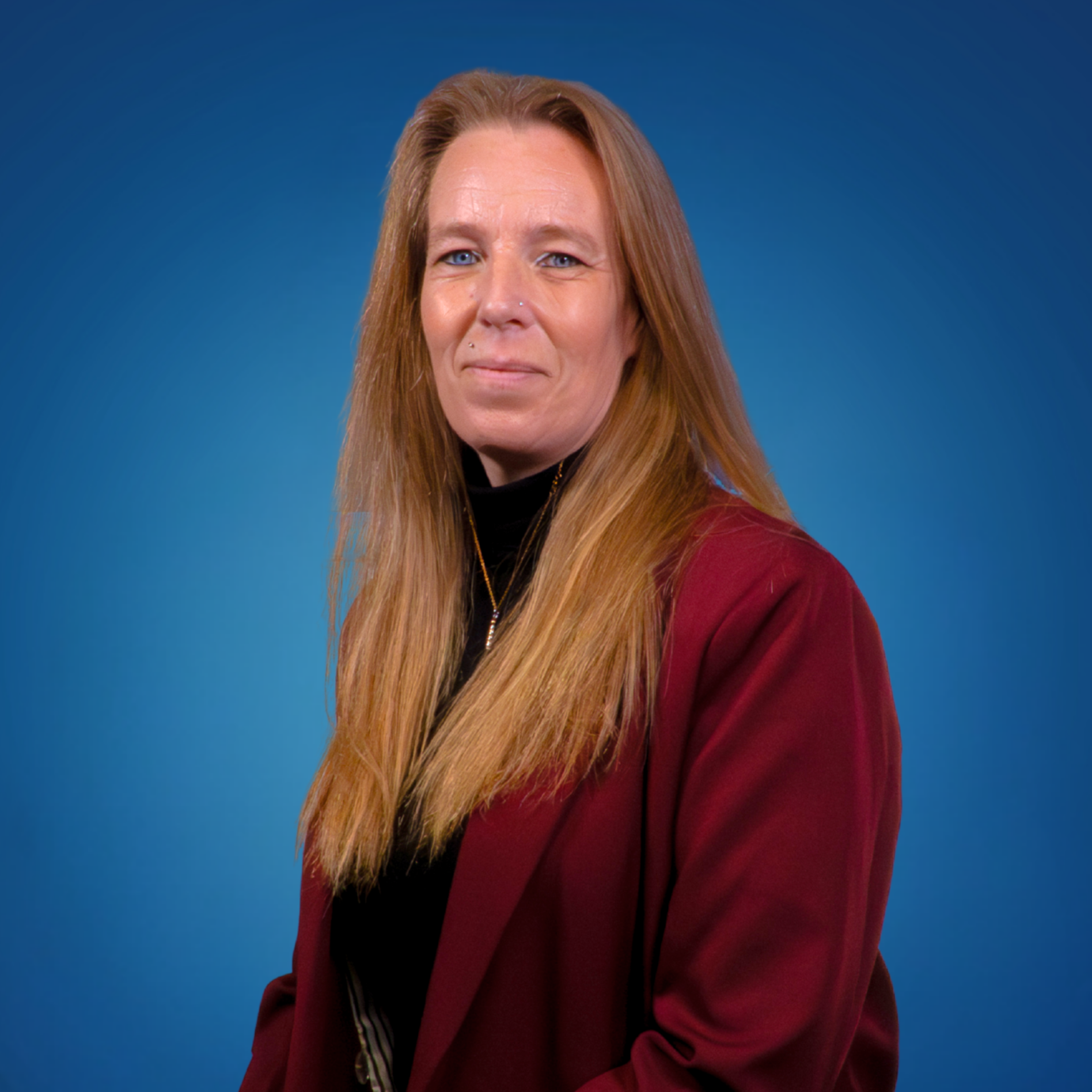 A woman with long reddish-blonde hair wearing a maroon blazer and black turtleneck stands against a blue background.
