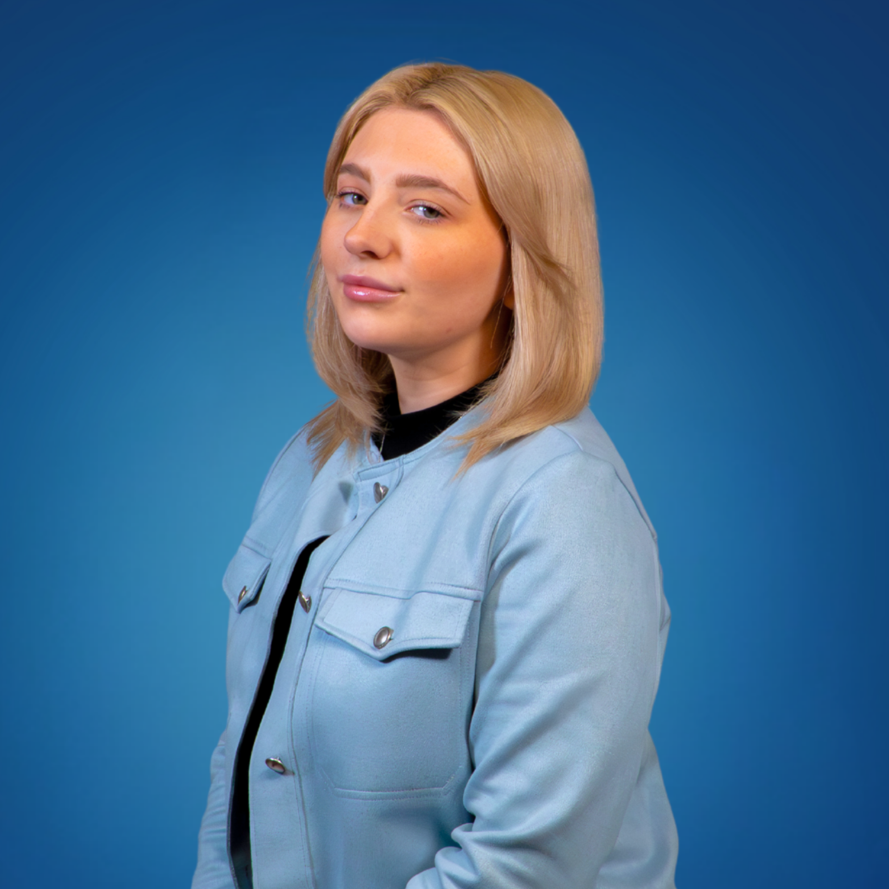 A young woman with blonde hair and fair skin, wearing a light blue jacket over a black top, posing against a solid blue background.