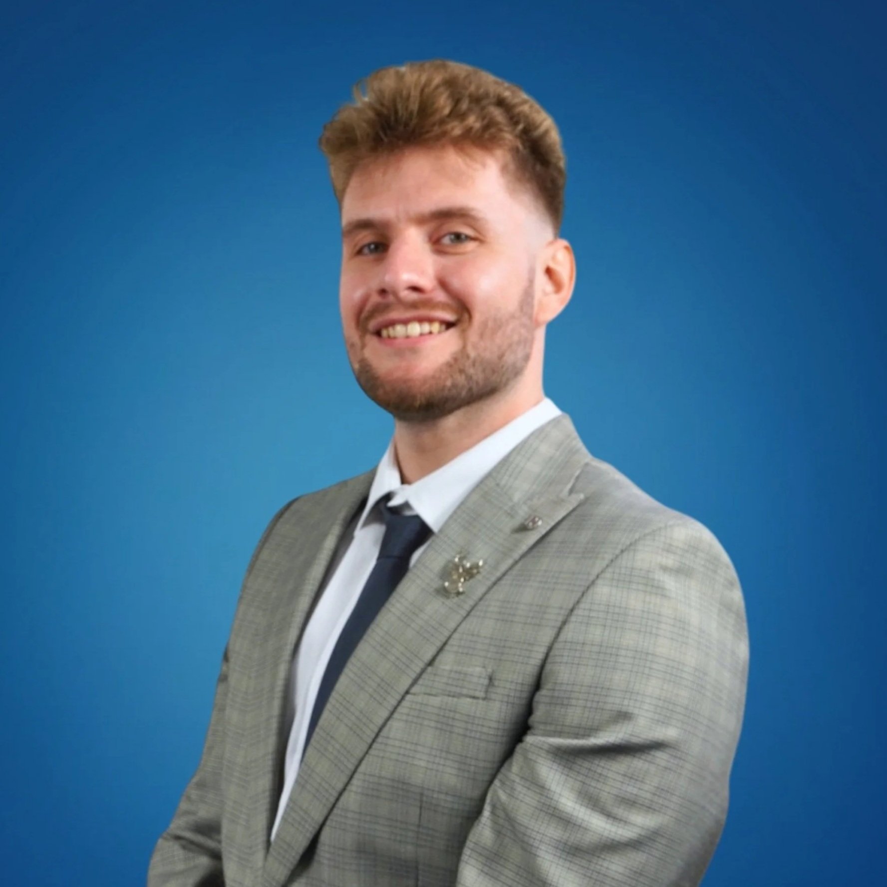 A young man with light brown hair styled upwards, smiling, wearing a light gray checkered suit jacket with a small pin on the lapel, a white dress shirt, and a dark tie, standing in front of a solid blue background.