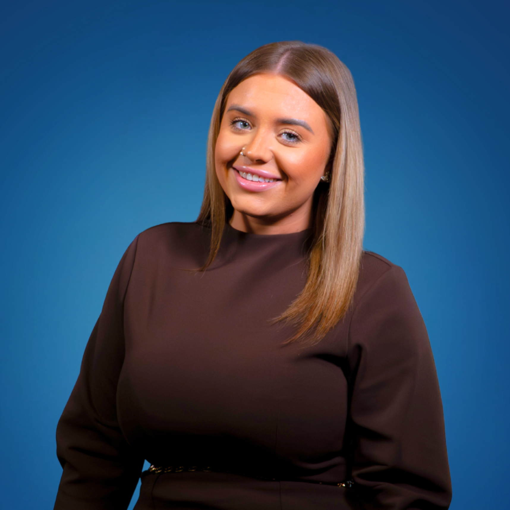 Smiling woman with straight brown hair, wearing a dark brown top, posing against a blue background.