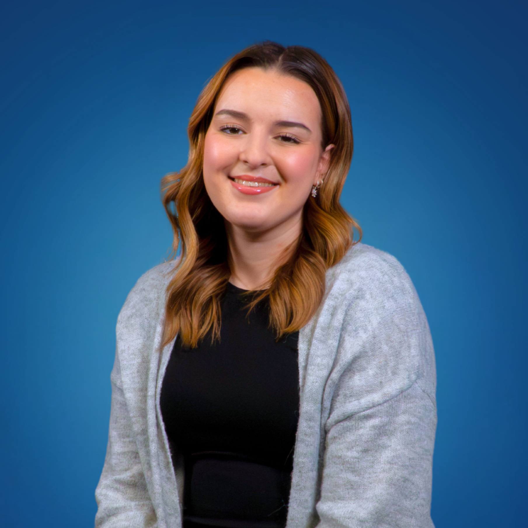 Portrait of a young woman with brown and blonde hair, wearing a gray cardigan over a black top, smiling against a blue background.