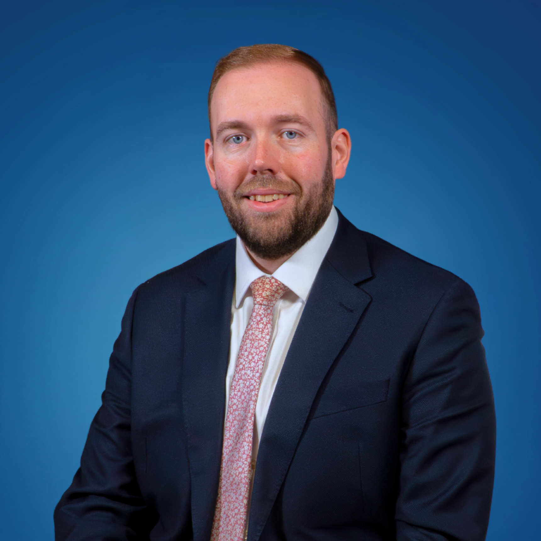 Professional headshot of a man in a dark suit with a white shirt and patterned pink tie, smiling, against a blue background.