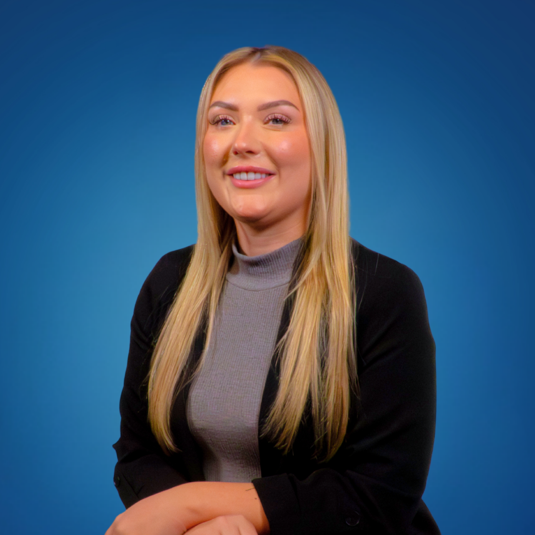A smiling blonde woman with long hair wearing a gray turtleneck and black blazer against a blue background.