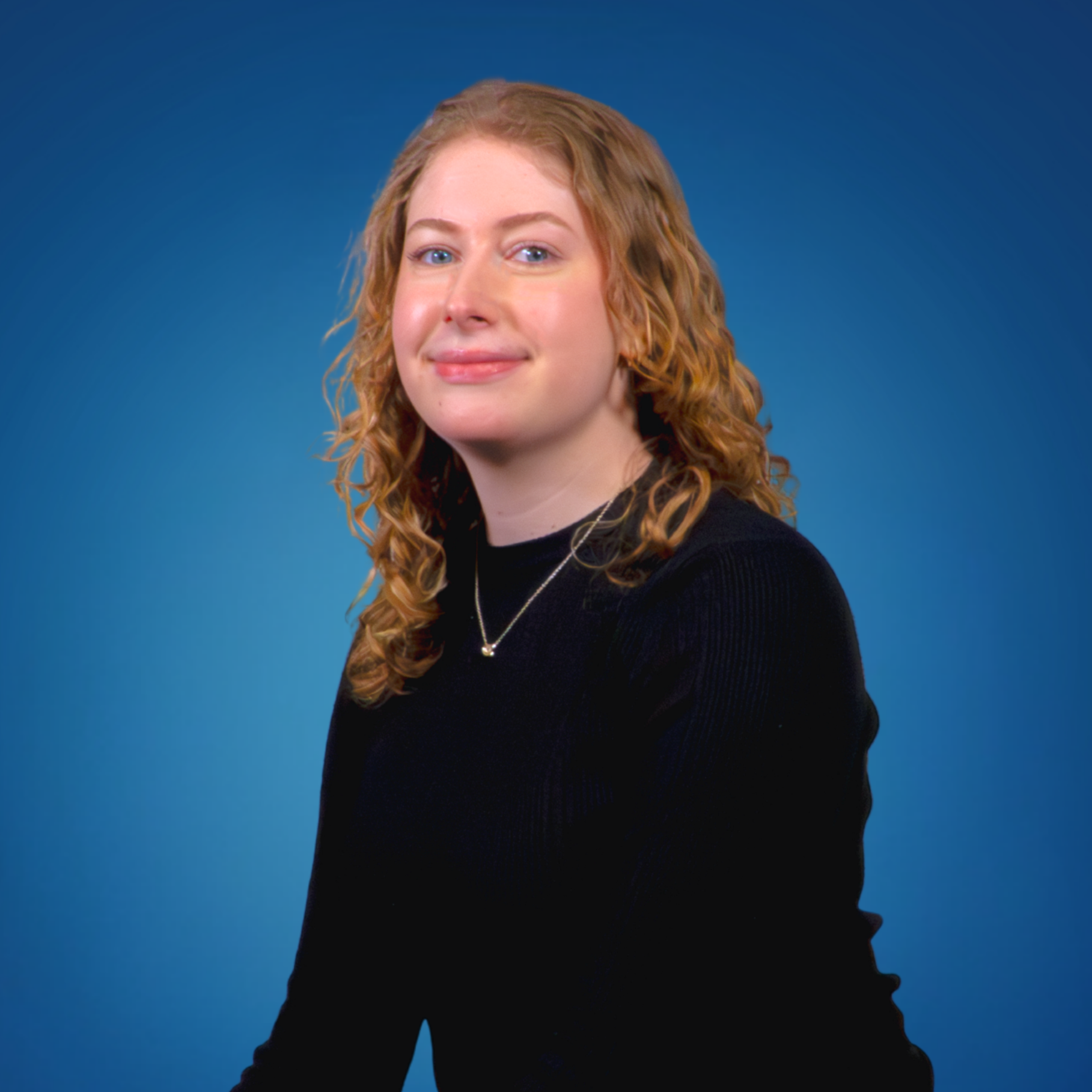 A young woman with red curly hair, wearing a black top and a necklace, smiling in front of a blue background.