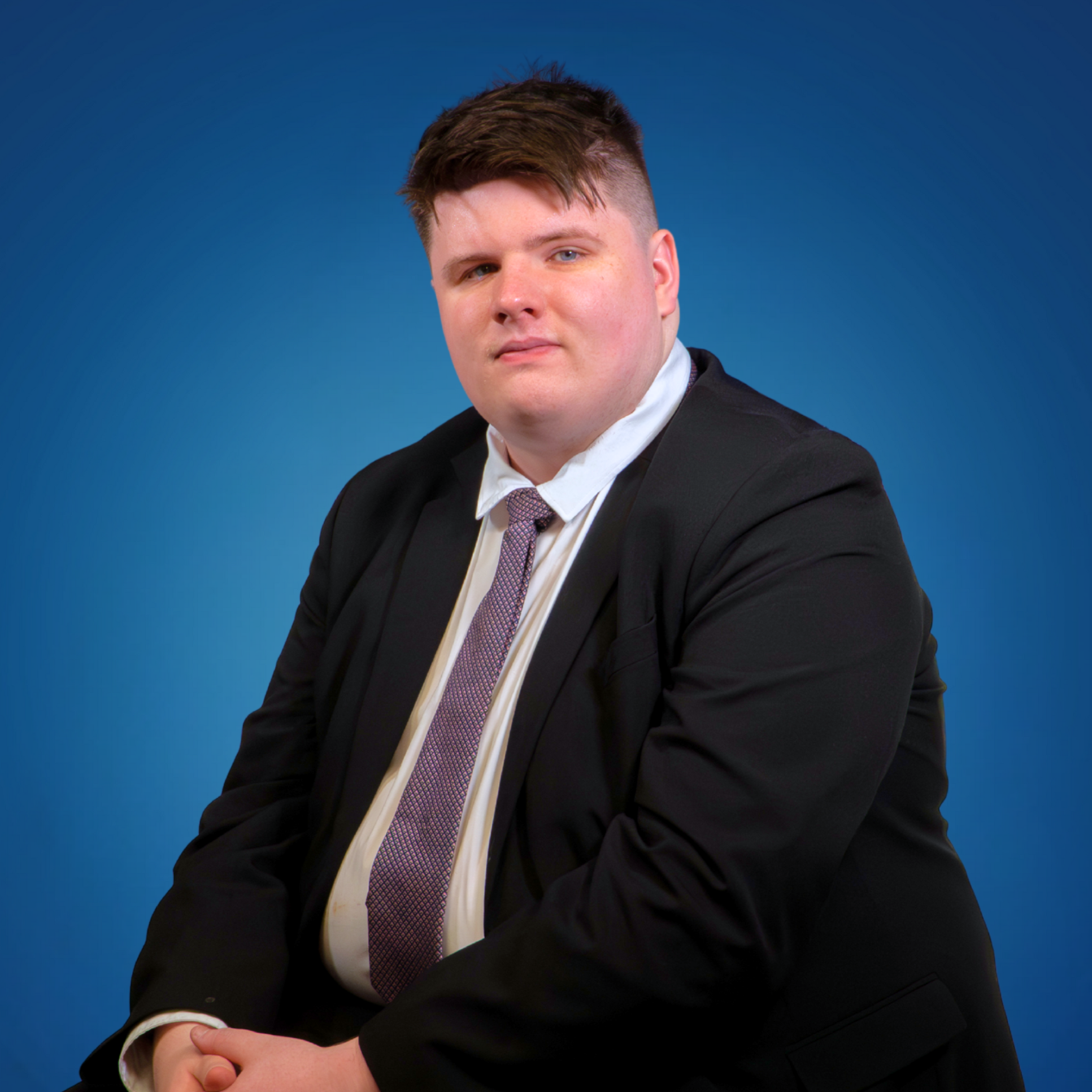 A man in a black suit with a white shirt and a patterned tie, sitting against a blue background.
