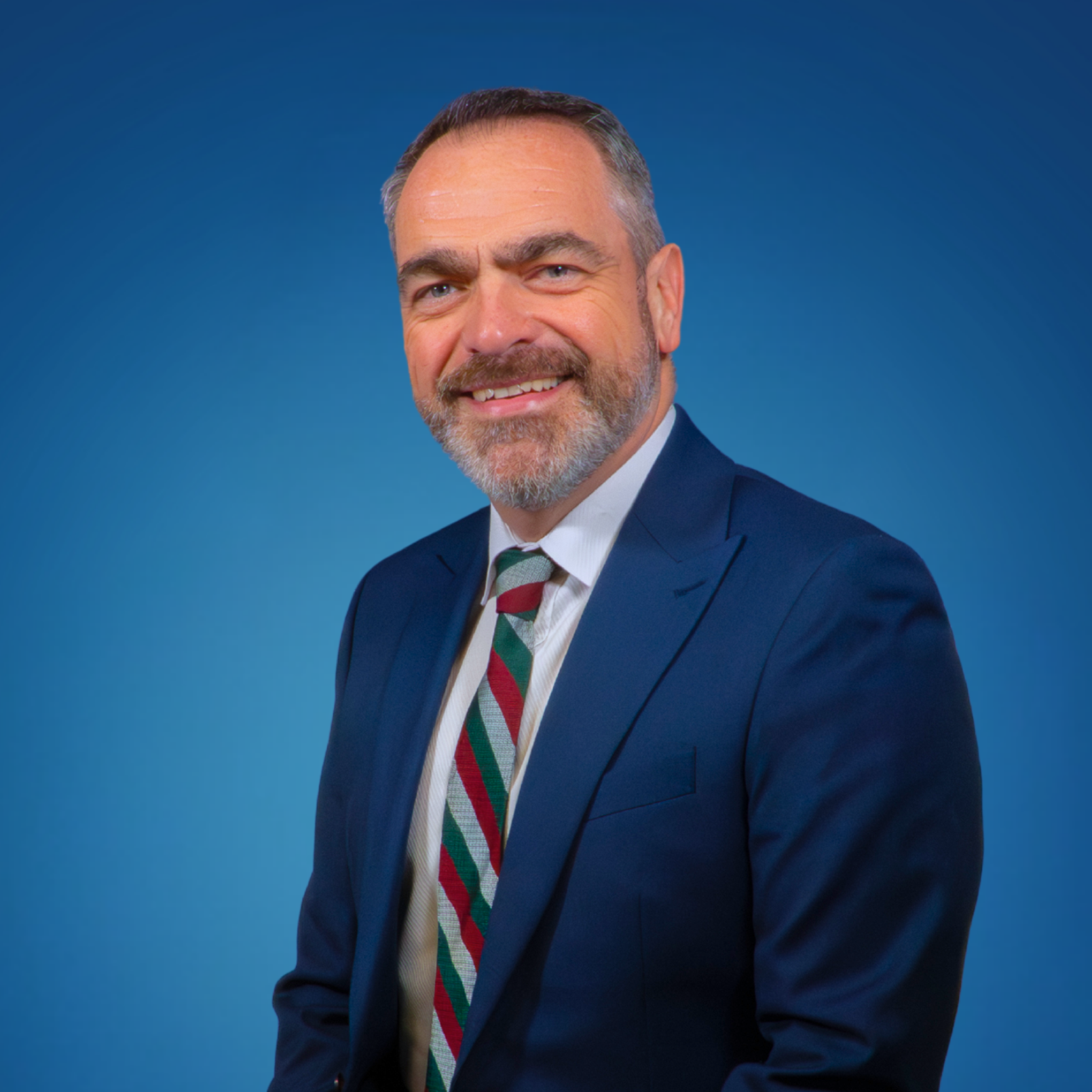 Professional portrait of a middle-aged man with a beard and mustache, wearing a dark blue suit, white shirt, and a striped holiday-themed tie, smiling against a blue background.