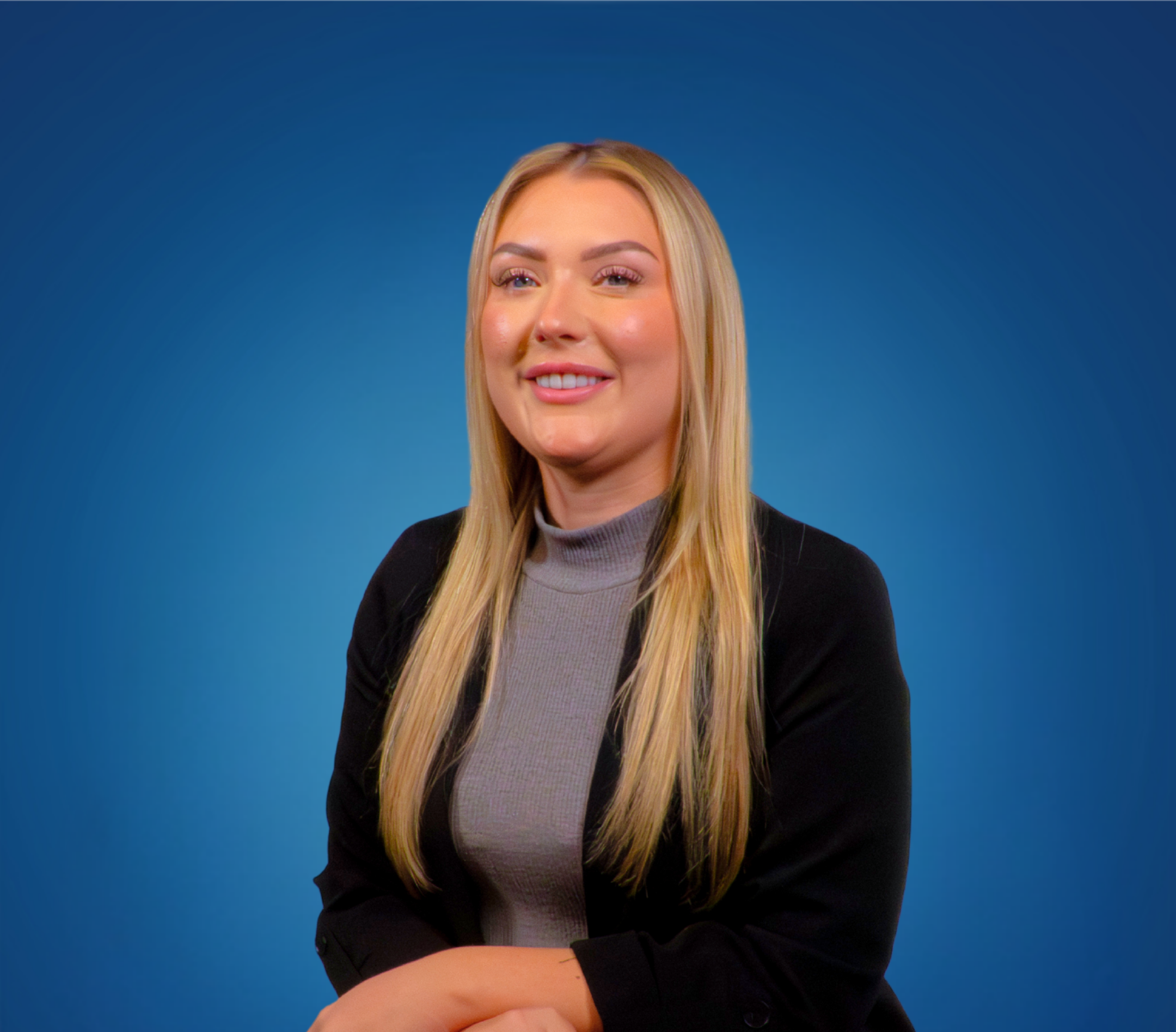 A blonde woman wearing a gray turtleneck and black blazer smiling against a blue gradient background.
