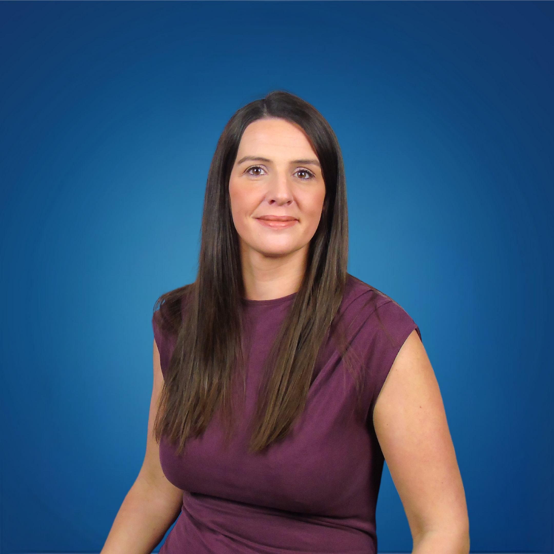 A woman with long brown hair and light skin, wearing a sleeveless purple top, is standing against a blue background.