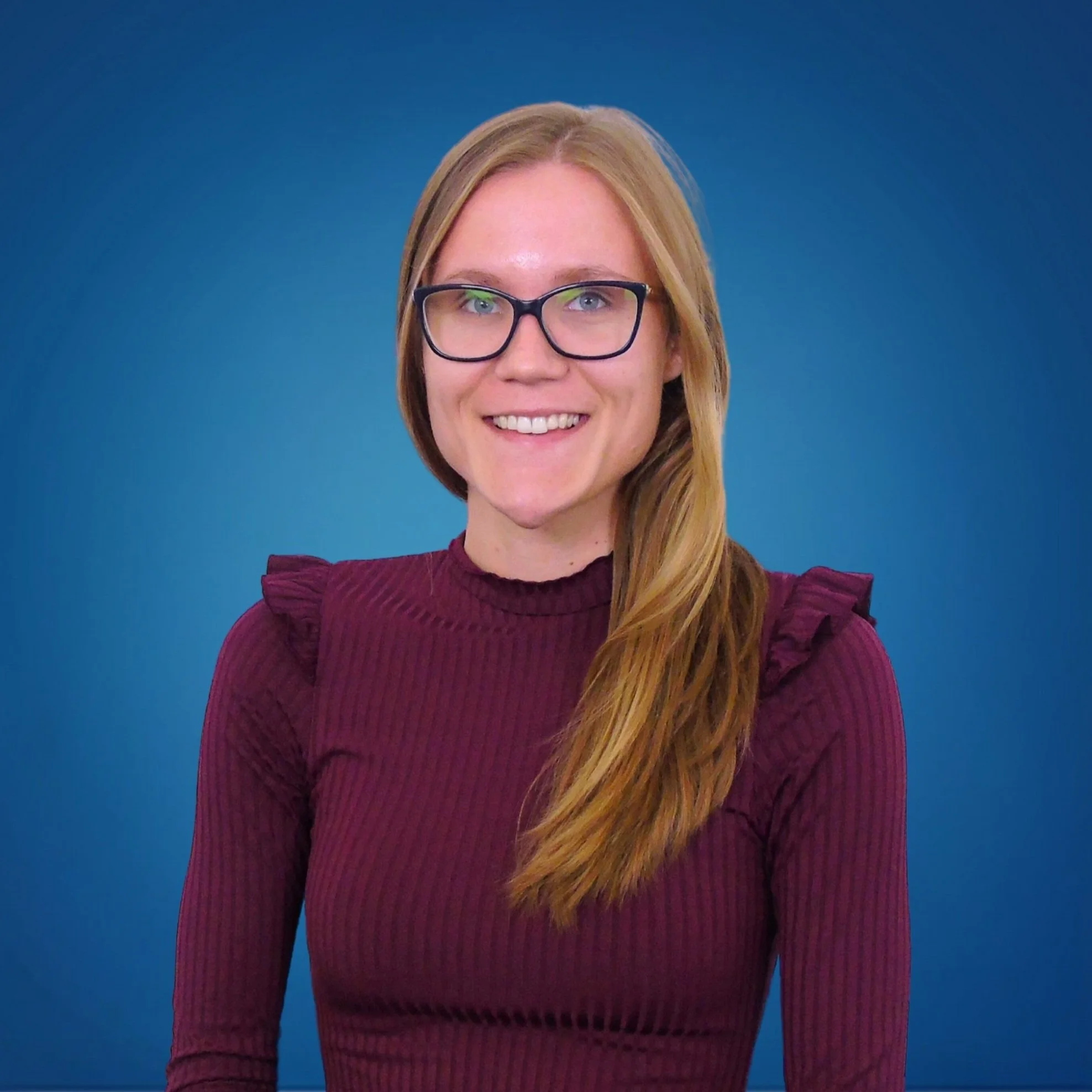 Portrait of a woman with long red hair and glasses, wearing a maroon top, smiling in front of a blue background.