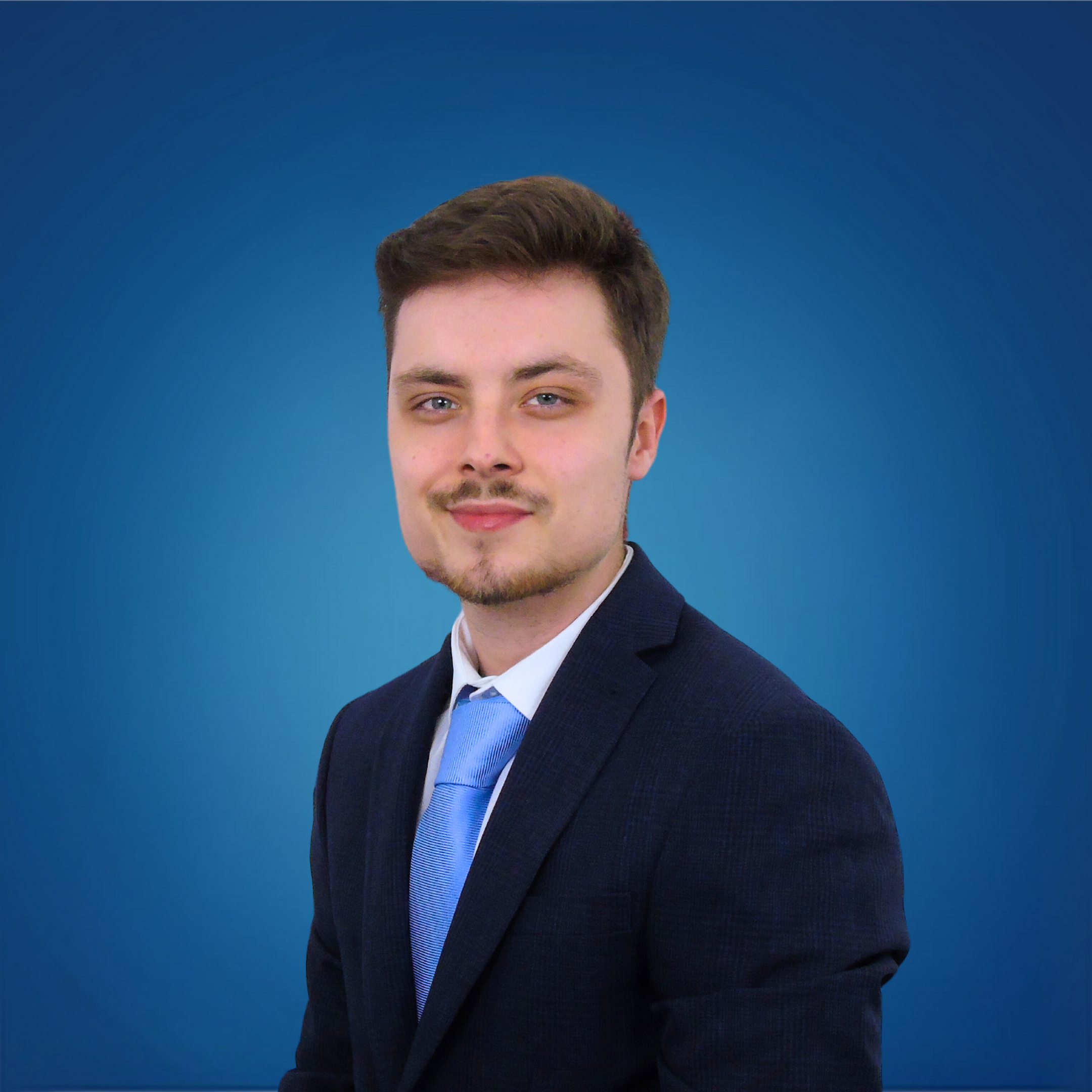 A young man with short brown hair, blue eyes, and light facial hair wearing a dark suit and light blue tie, standing against a solid blue background.