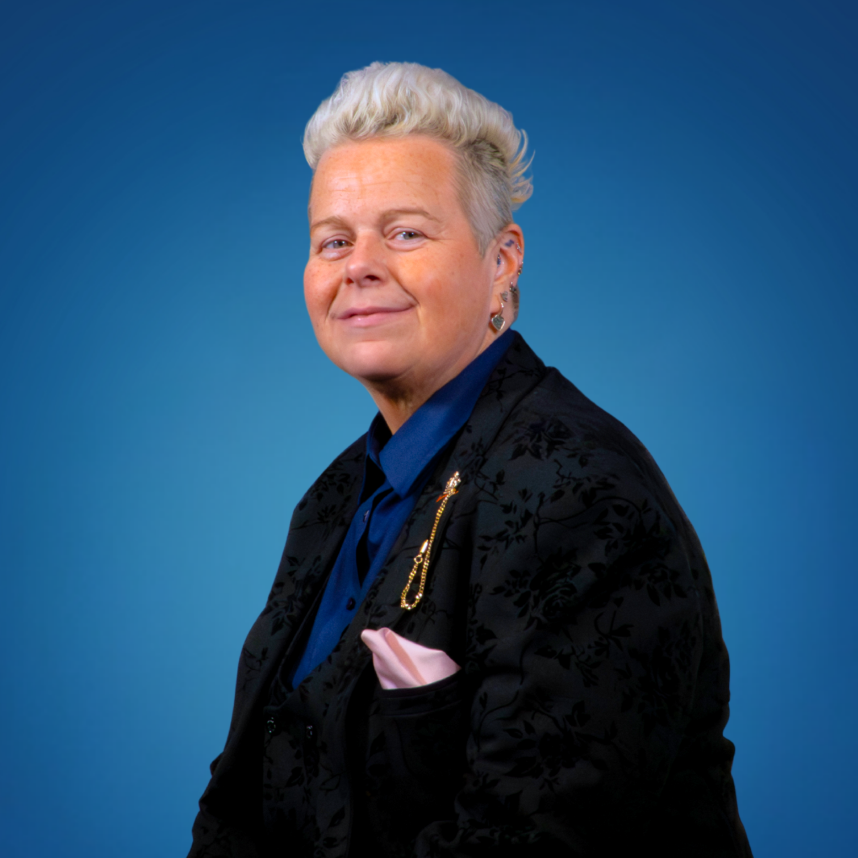 Portrait of a woman with short, curly platinum blonde hair, wearing a dark textured blazer, a blue collared shirt, and jewelry, against a gradient blue background.