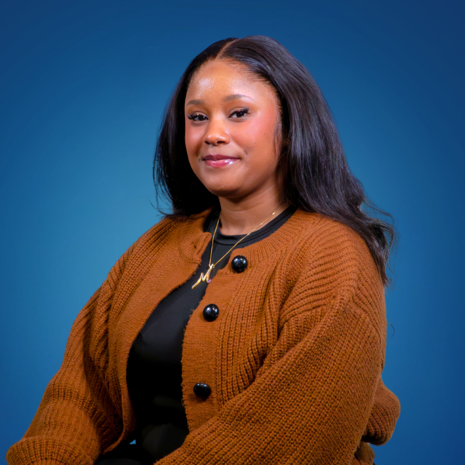 Portrait of a young woman with black hair and a warm smile, wearing a brown jacket with black buttons, a black top, and a gold necklace with a pendant shaped like a letter 'M', against a blue background.