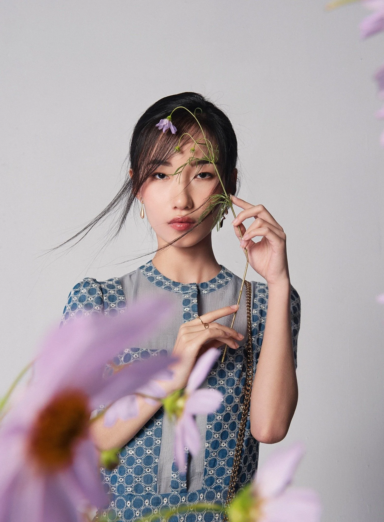 Retrato de una mujer con cabello negro, sosteniendo una flor y rodeada de flores lilas, en un fondo gris claro.