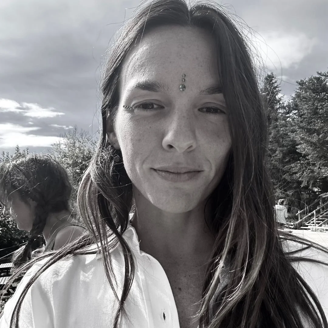 Retrato en blanco y negro de una mujer con cabello largo y oscuro, con un piercing en el ojo izquierdo y joyas en la cara, al aire libre con árboles y cielo nublado al fondo.