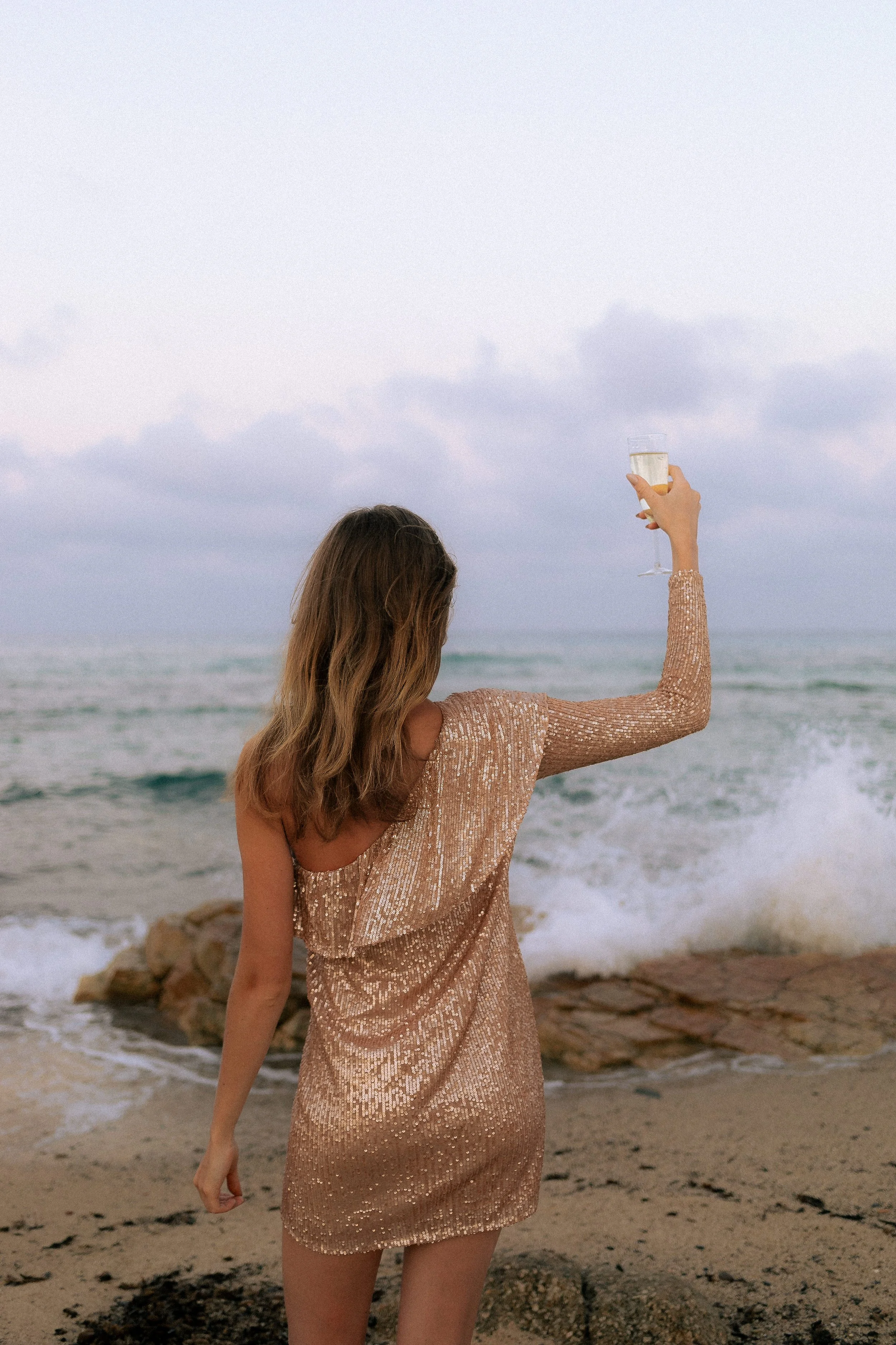 Mujer con vestido dorado de lentejuelas, de espaldas, sosteniendo una copa de champagne frente al mar.
