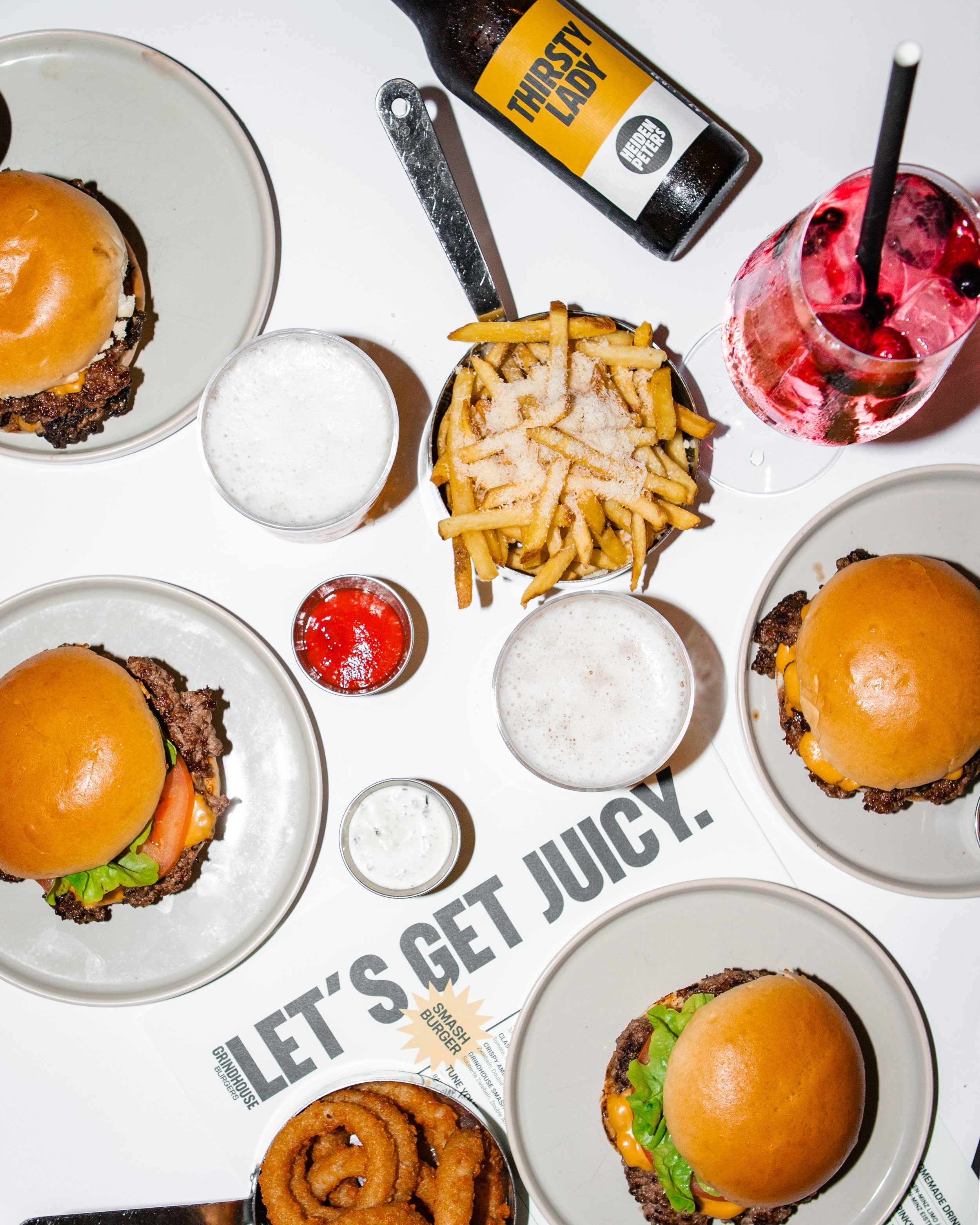 Mehrere Hamburger auf weißen Tellern, Pommes frites mit Salz, Zwiebelringe, verschiedene Getränke, inklusive Cola und alkoholfreie Getränke, auf einem Tisch mit einem Werbeaufdruck, der sagt 'Let's get juicy'.