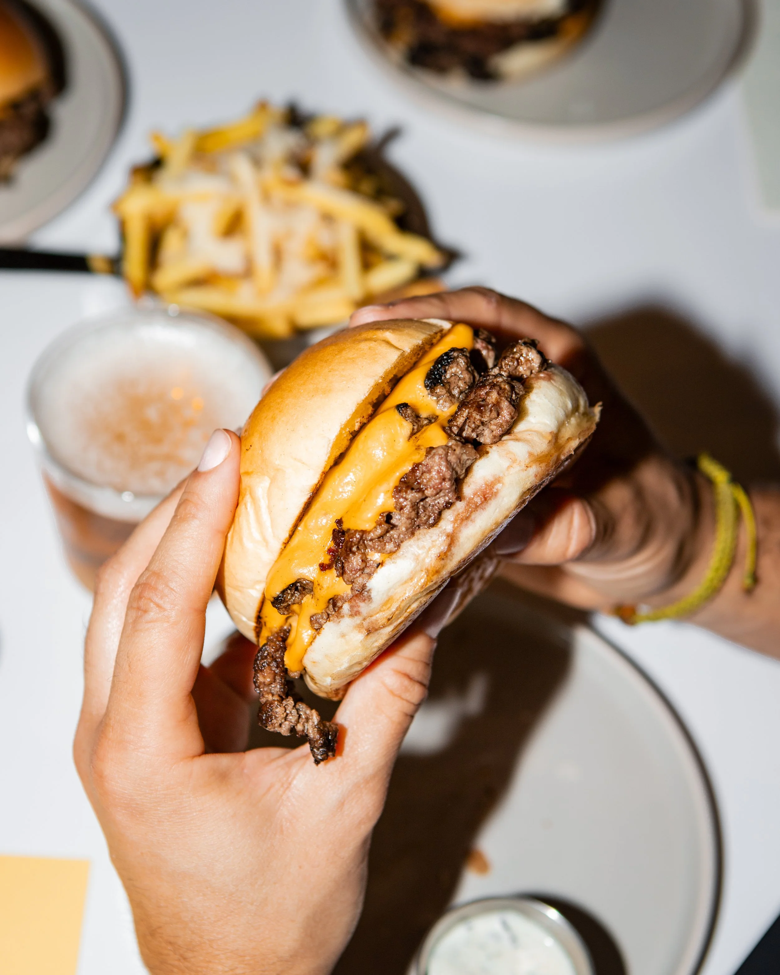 Person hält einen Cheeseburger mit Rindfleisch, Käse und Brötchen, im Hintergrund Pommes und Getränke auf einem Tisch, in Berlin Prenzlauer Berg. Beef ist von Weiderindern. Beef ist Halal. Auch als vegetarischer Burger zu bestellen.