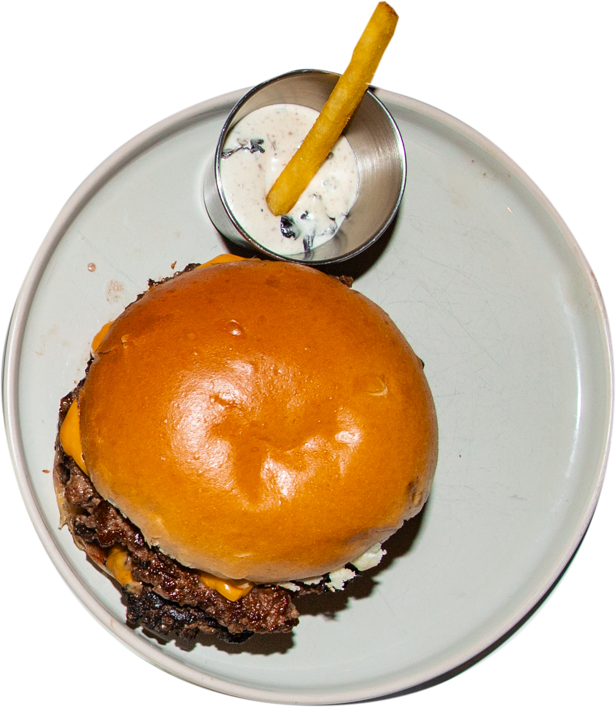 Ein Hamburger mit Rindfleischpatty, Käse und Brötchen, dazu eine kleine Schale mit Soße und einem Kartoffelstab auf einem runden weißen Teller.
