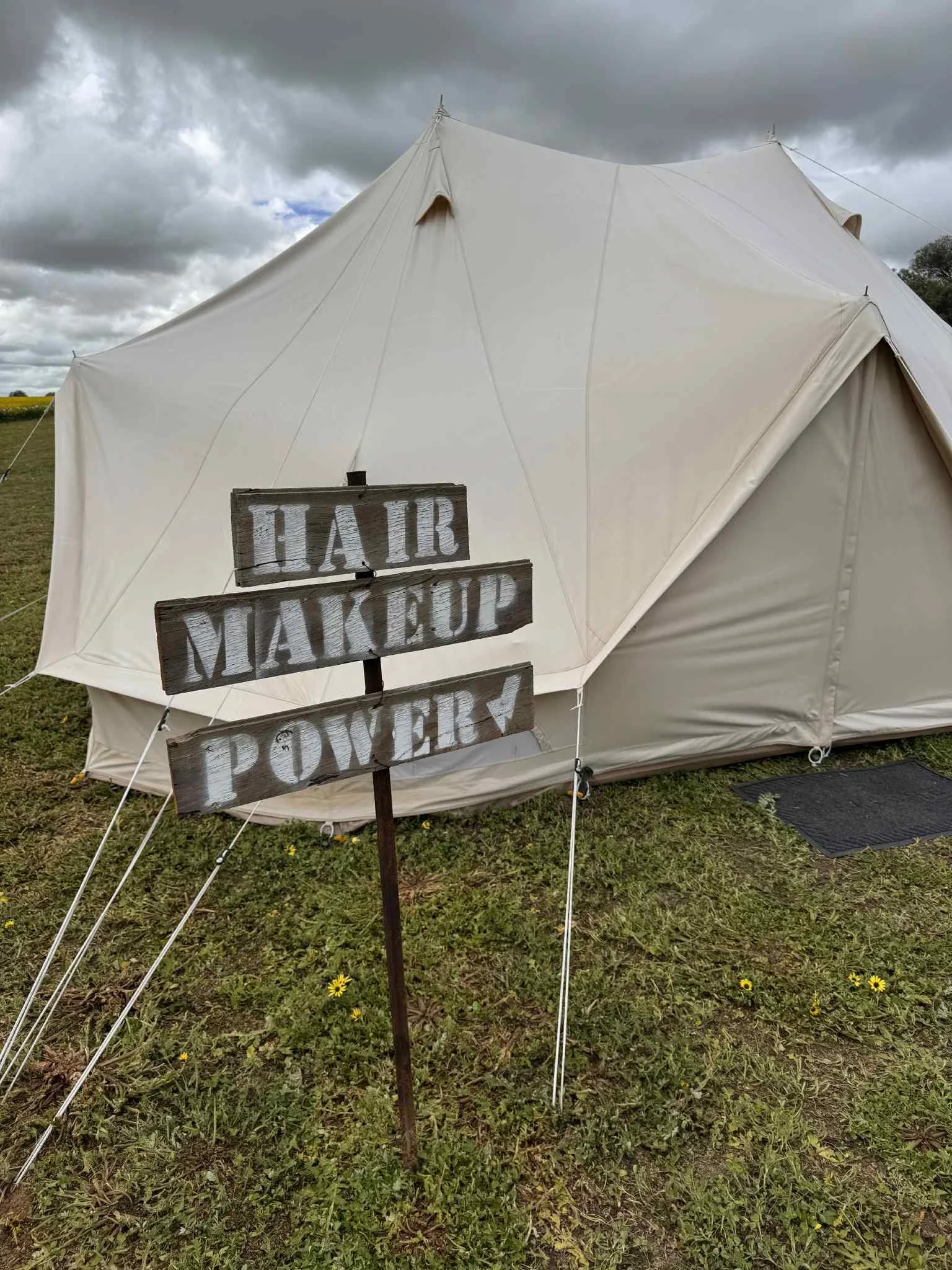 A white fabric tent set up outdoors on a grassy field under a cloudy sky, with a wooden sign in front that reads "HAIR MAKEUP DELIVERY."