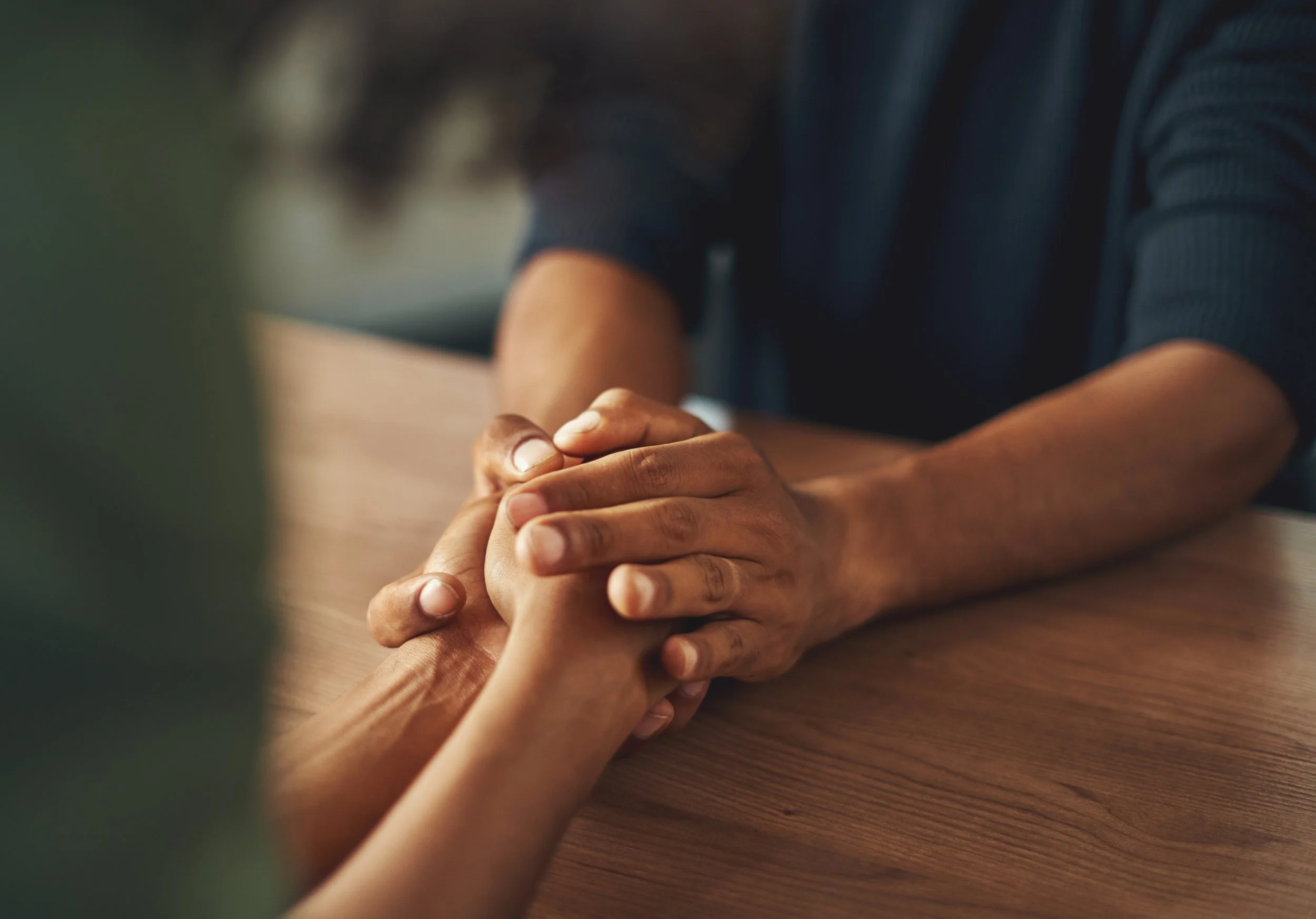 Two people holding hands on a wooden table, with one person's hands gently grasping the other's.