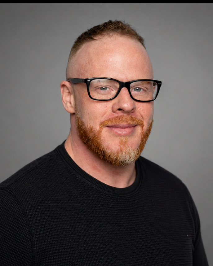 A man with red hair and beard wearing black glasses and a gray t-shirt standing outdoors with blurred trees and sunlight in the background.