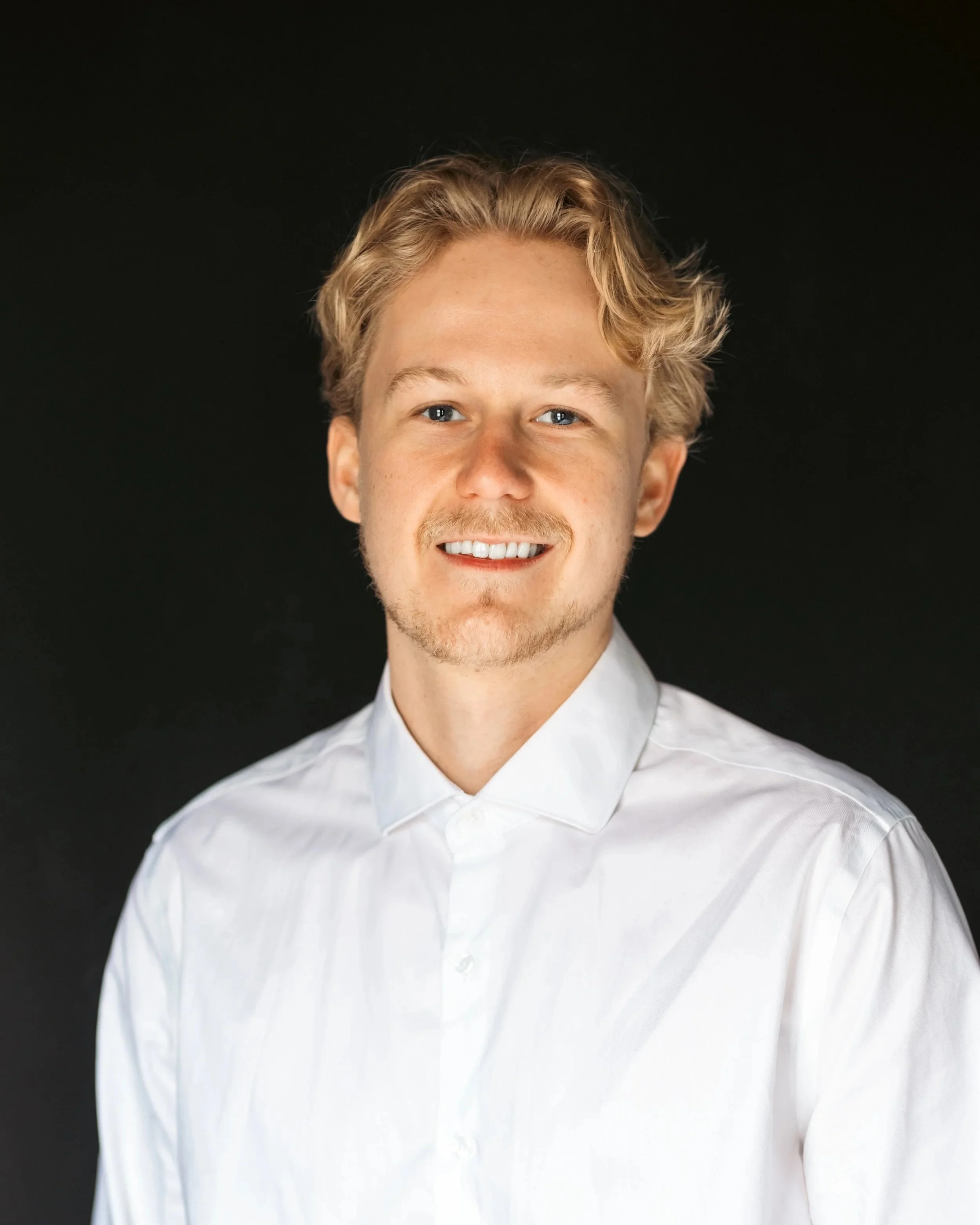 A young man with blonde curly hair smiling, wearing a black collared shirt and a white undershirt, against a plain light-colored background.