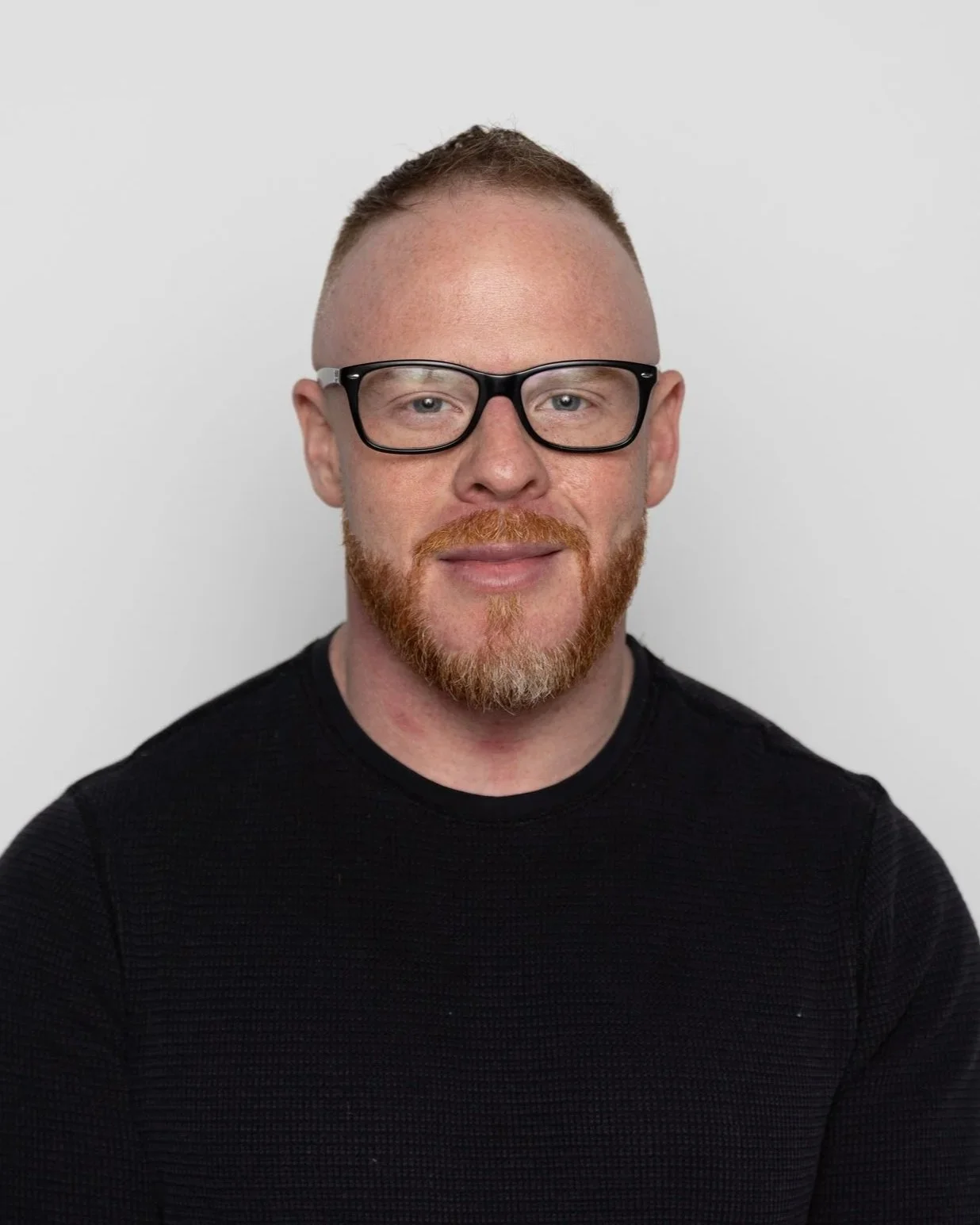 A man with red hair and beard wearing black glasses and a gray t-shirt standing outdoors with blurred trees and sunlight in the background.
