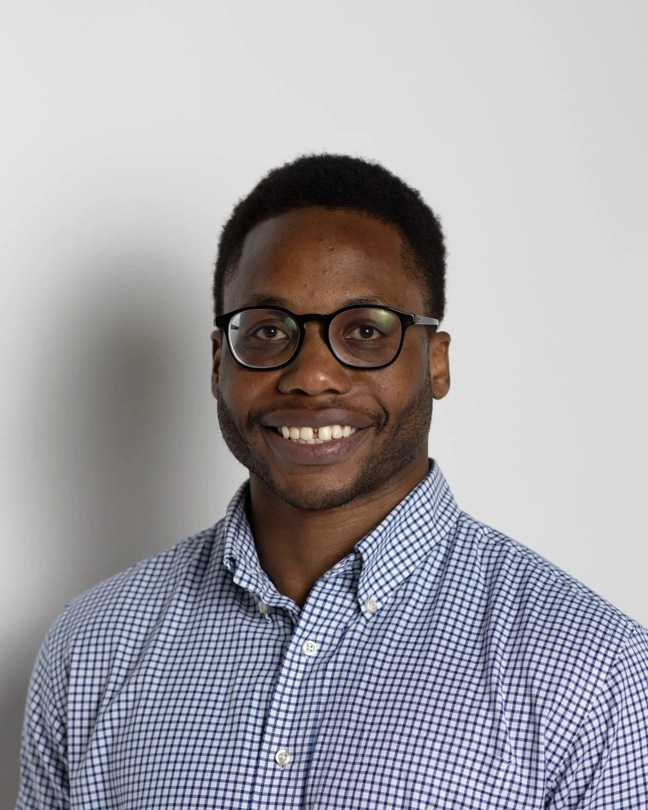 A man with short black hair, glasses, and a beard, smiling, wearing a blue and white checkered collared shirt, standing against a plain white wall.