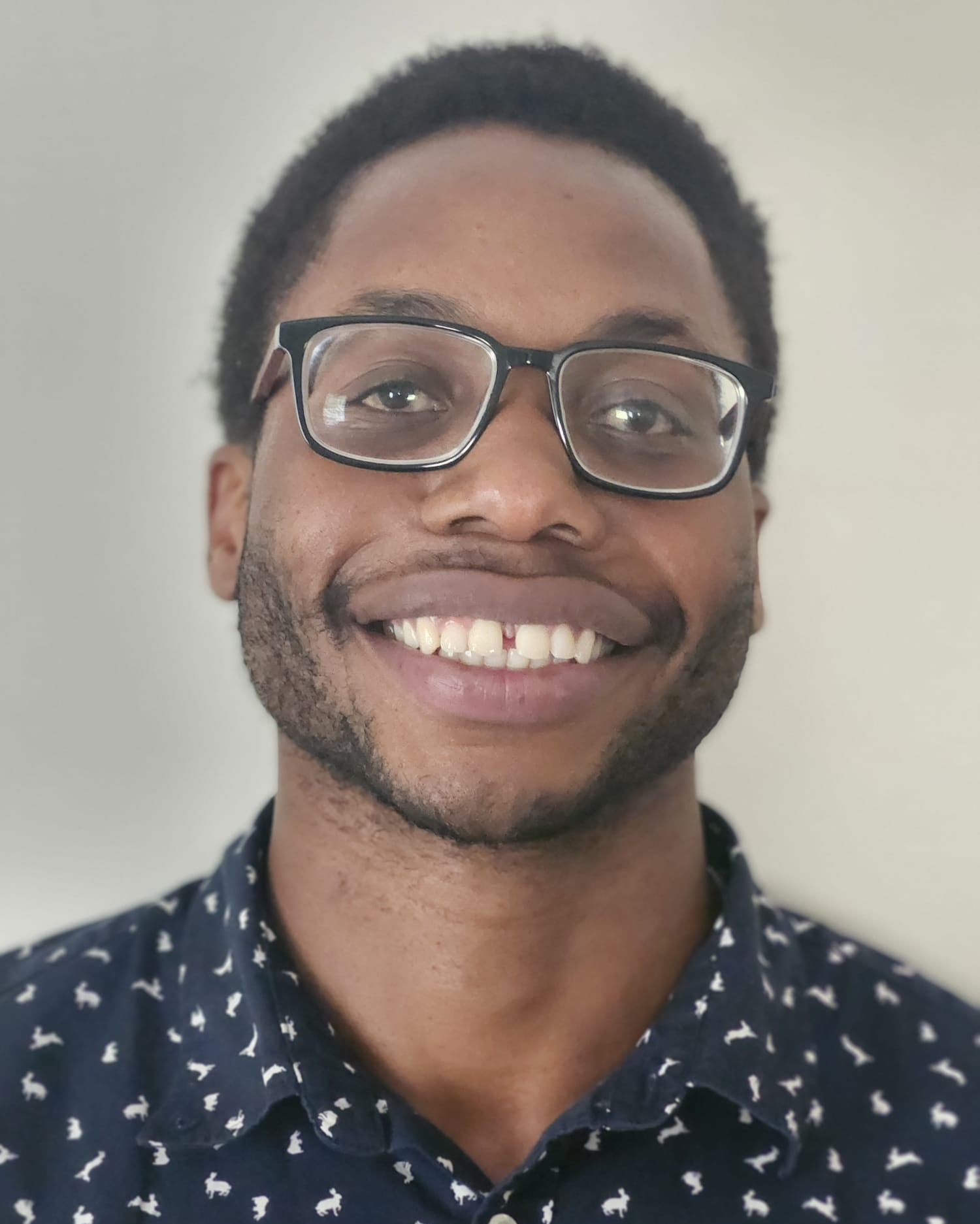 Close-up of a smiling man with glasses, short curly hair, and a patterned shirt against a plain background.