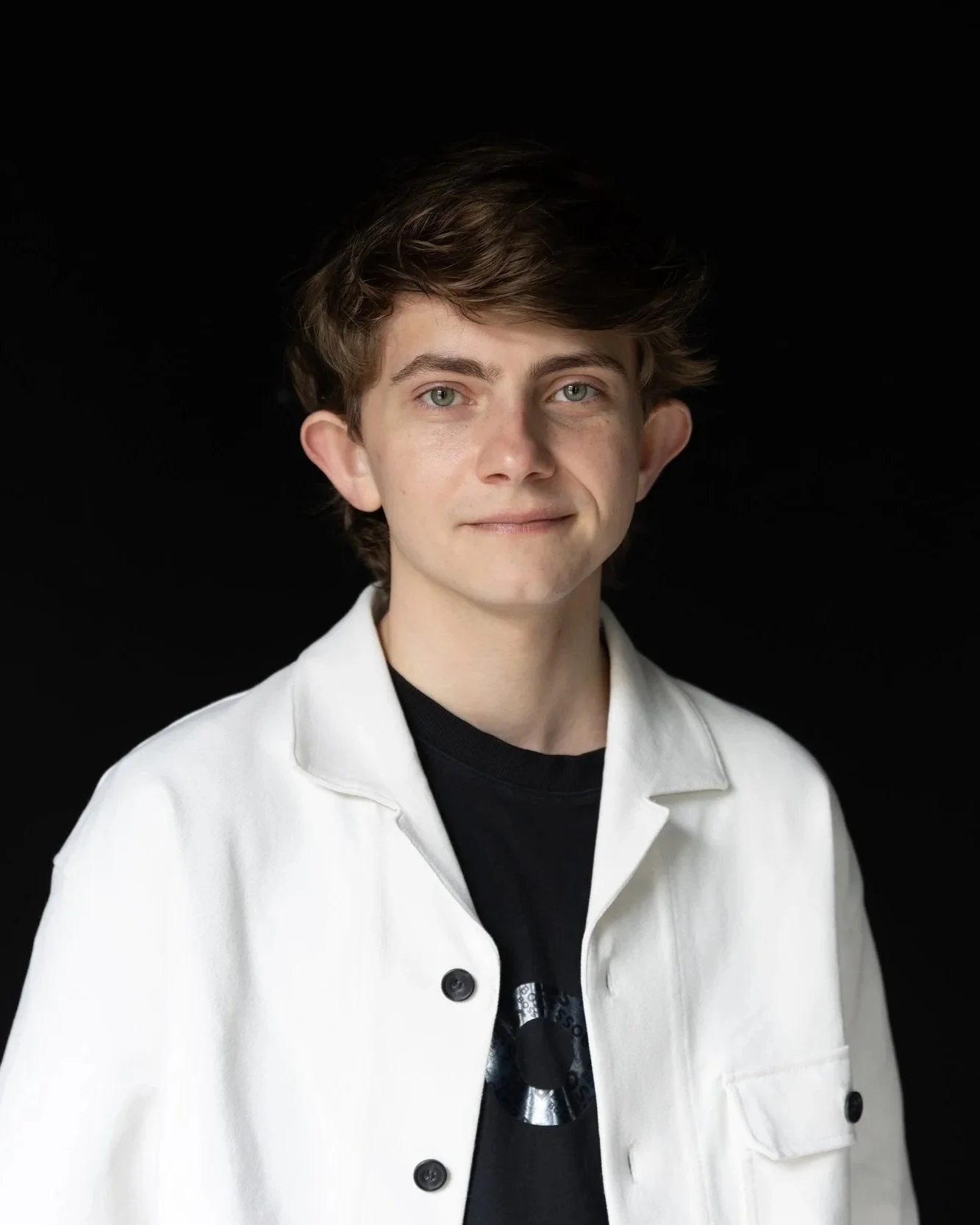 Portrait of a young man with light skin, wavy brown hair, and green eyes wearing a white jacket over a black shirt, standing against a black background.