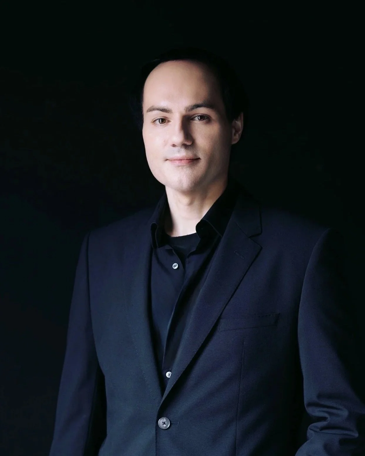 Professional headshot of a smiling young man with dark hair, wearing a dark suit jacket over a blue shirt, against a gray background.