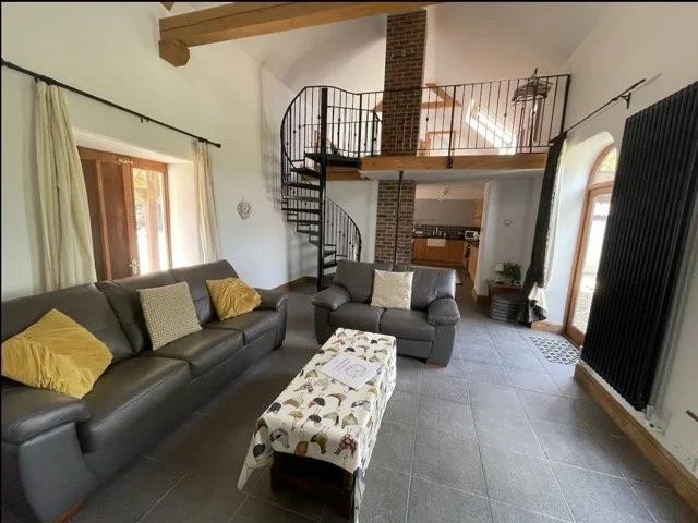 Cozy living room with gray sofas, a patterned coffee table, spiral staircase, and exposed wooden beams. There is a mezzanine with railings and a view of the kitchen in the background. Large windows allow natural light into the space.