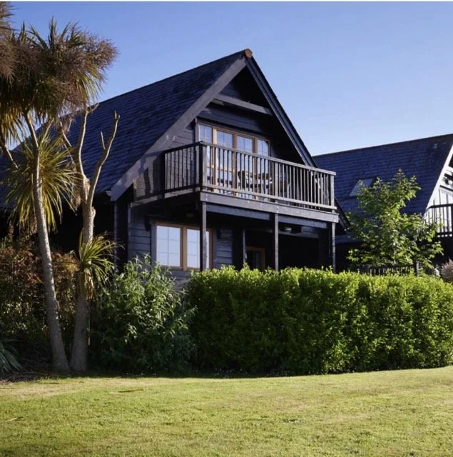 A wooden house with a balcony, surrounded by greenery and palm trees, on a sunny day.