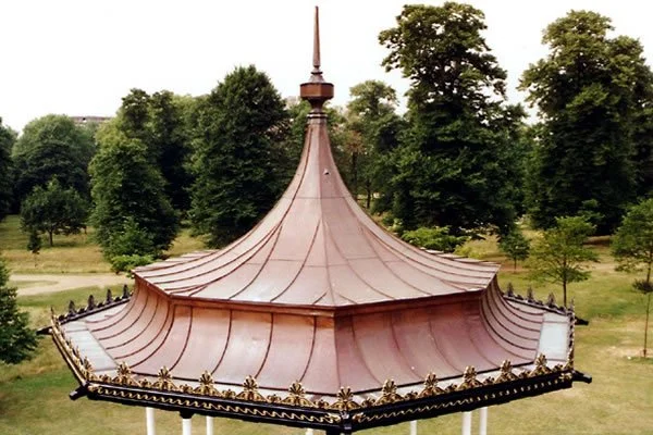 A pavilion with a pointed, curved copper roof surrounded by green trees and open grassy area.