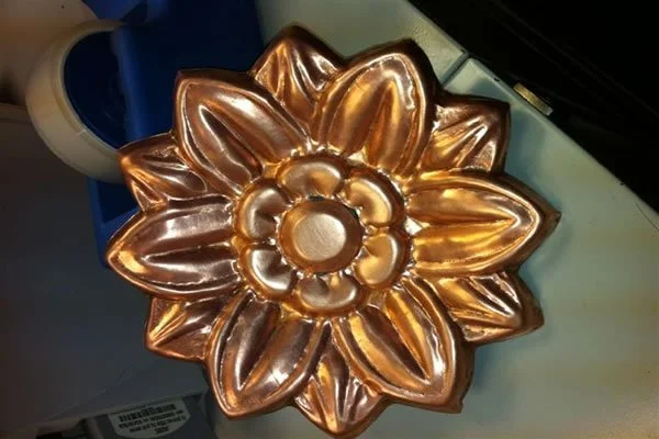 A metallic gold flower-shaped ornament with a circular center, placed on a surface near a roll of tape and a container.