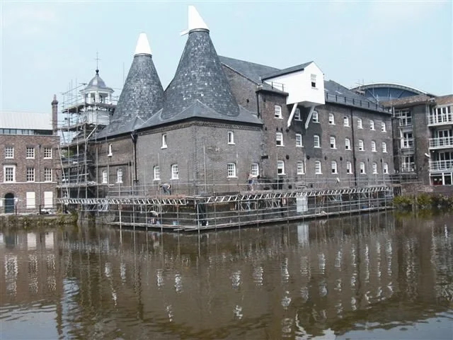 Historic brick building with conical roofs, surrounded by water with scaffolding and a small white projection on the side.