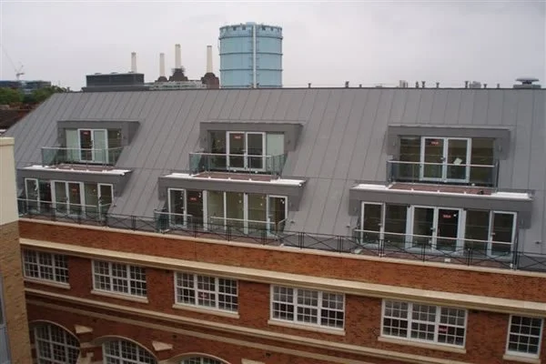 The image shows a rooftop with balconies, part of a brick building with large windows, and an industrial area with smokestacks and a water tower in the background.