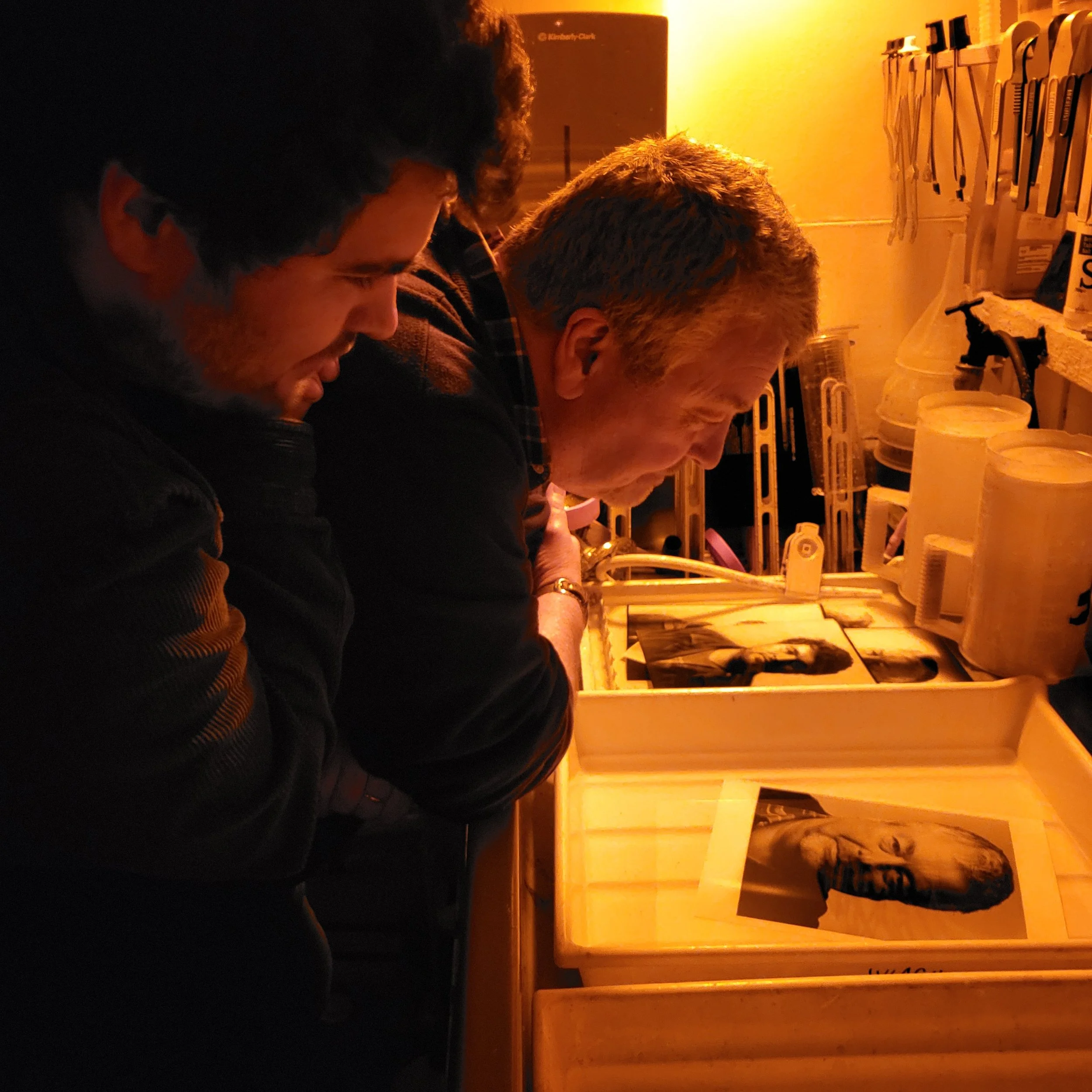 Two men are closely examining photographs at a darkroom sink in a dimly lit room, surrounded by photo development supplies.