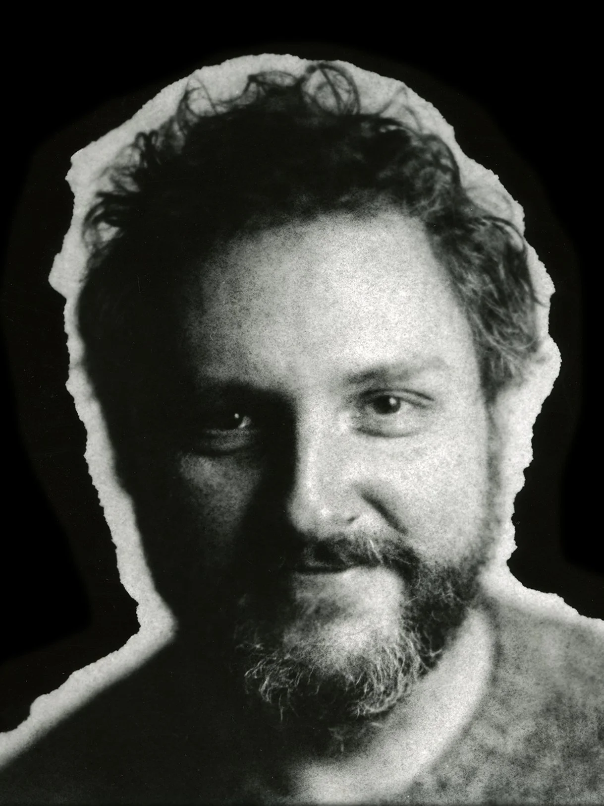 Black and white photo of a man's face with a beard and curly hair, torn edges around the photo.