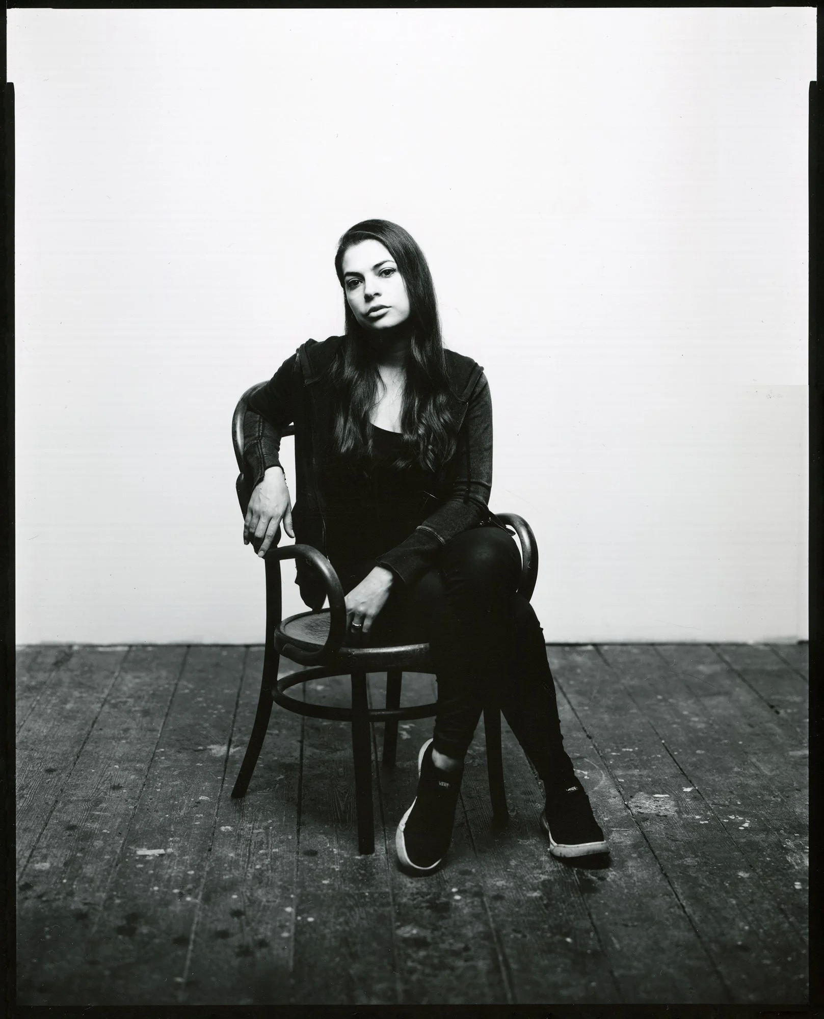 Black and white photo of a young woman sitting on a wooden chair against a blank wall. She has long, dark hair and is wearing dark clothing, including a hoodie, pants, and sneakers.